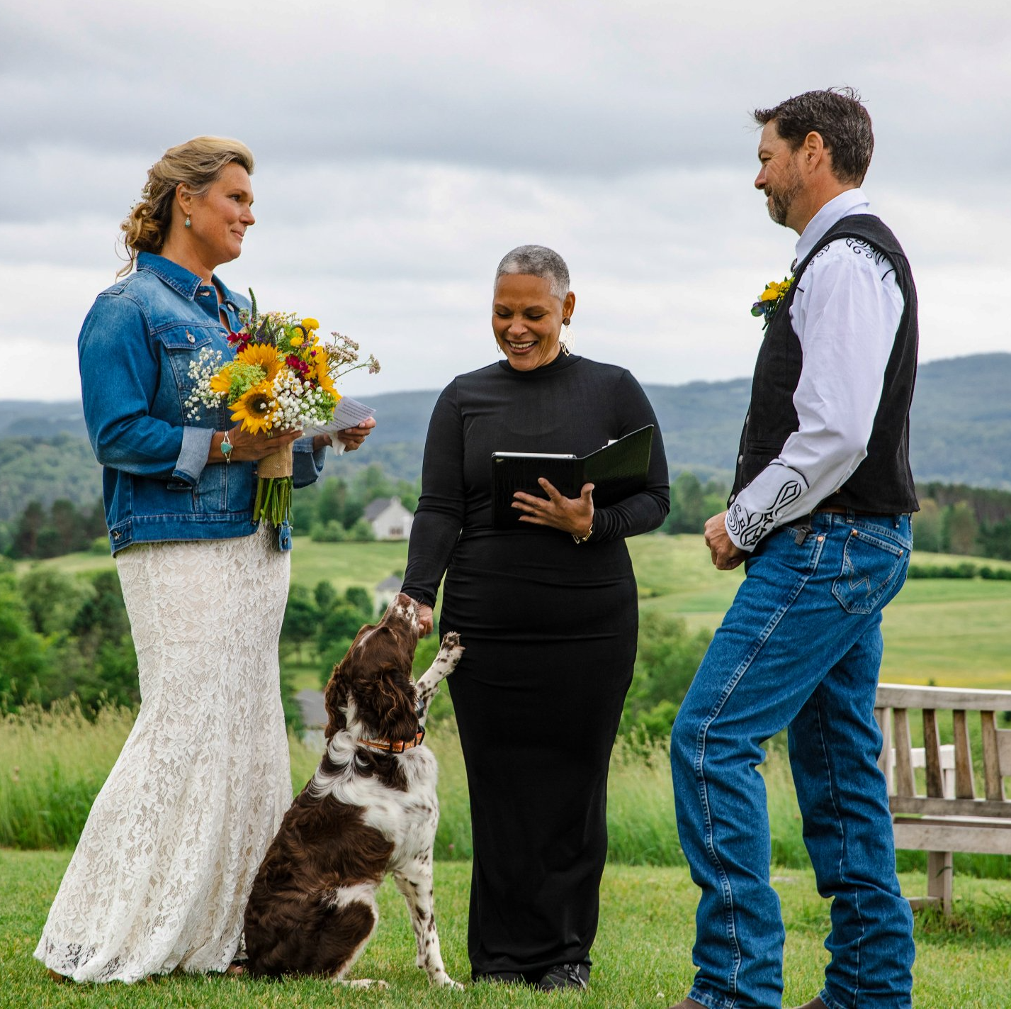 A bride and groom are standing in a field with their dog.