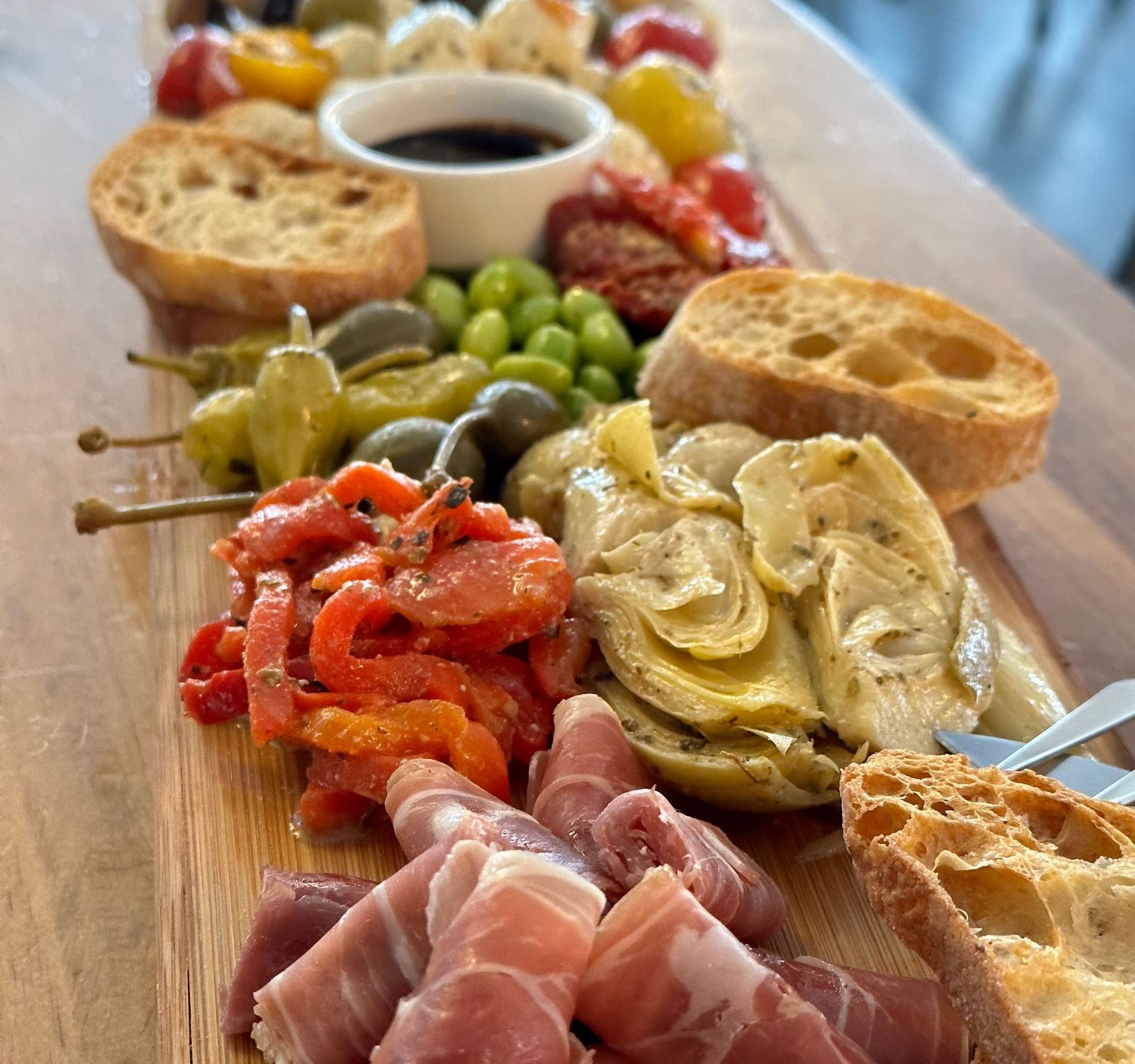 Closeup of antipasto or charcuterie board, including artichokes, red peppers, bread, cured meats, cheese, and tomatoes.