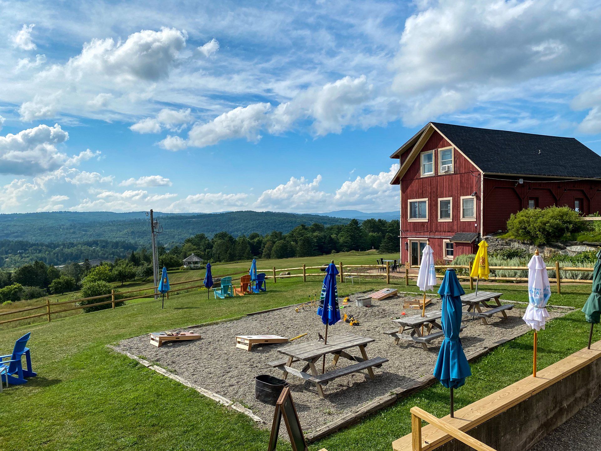 A red barn is sitting on top of a grassy hill next to a picnic area with tables and umbrellas.