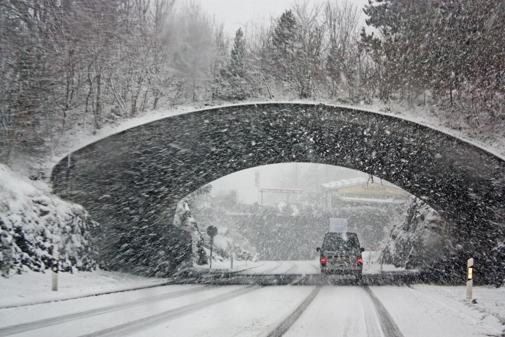 A truck driving on snow covered roads, while it is snowing, under a bridge