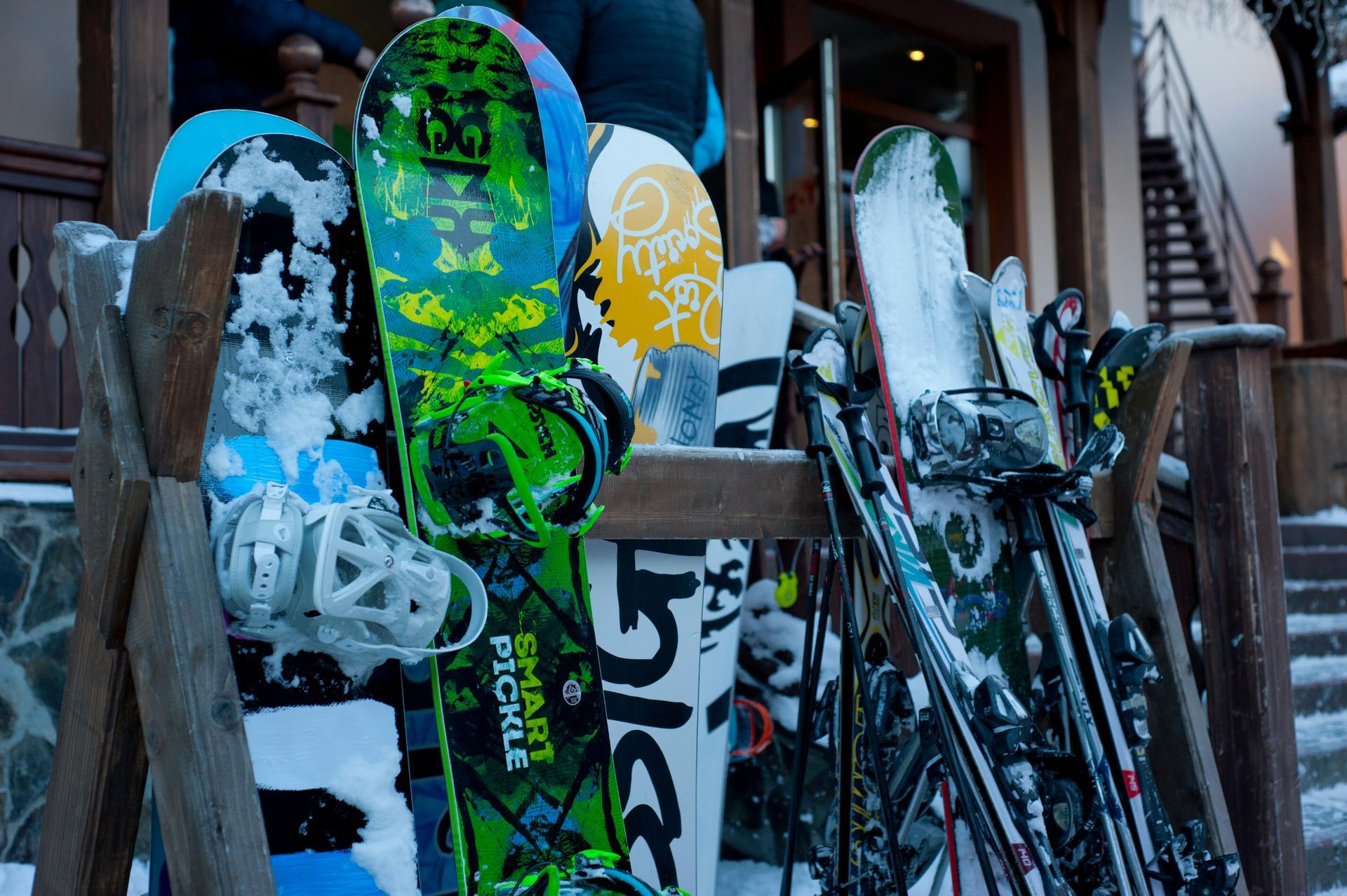 Snowboards and skis in a wooden rack, covered in snow, outside a building.