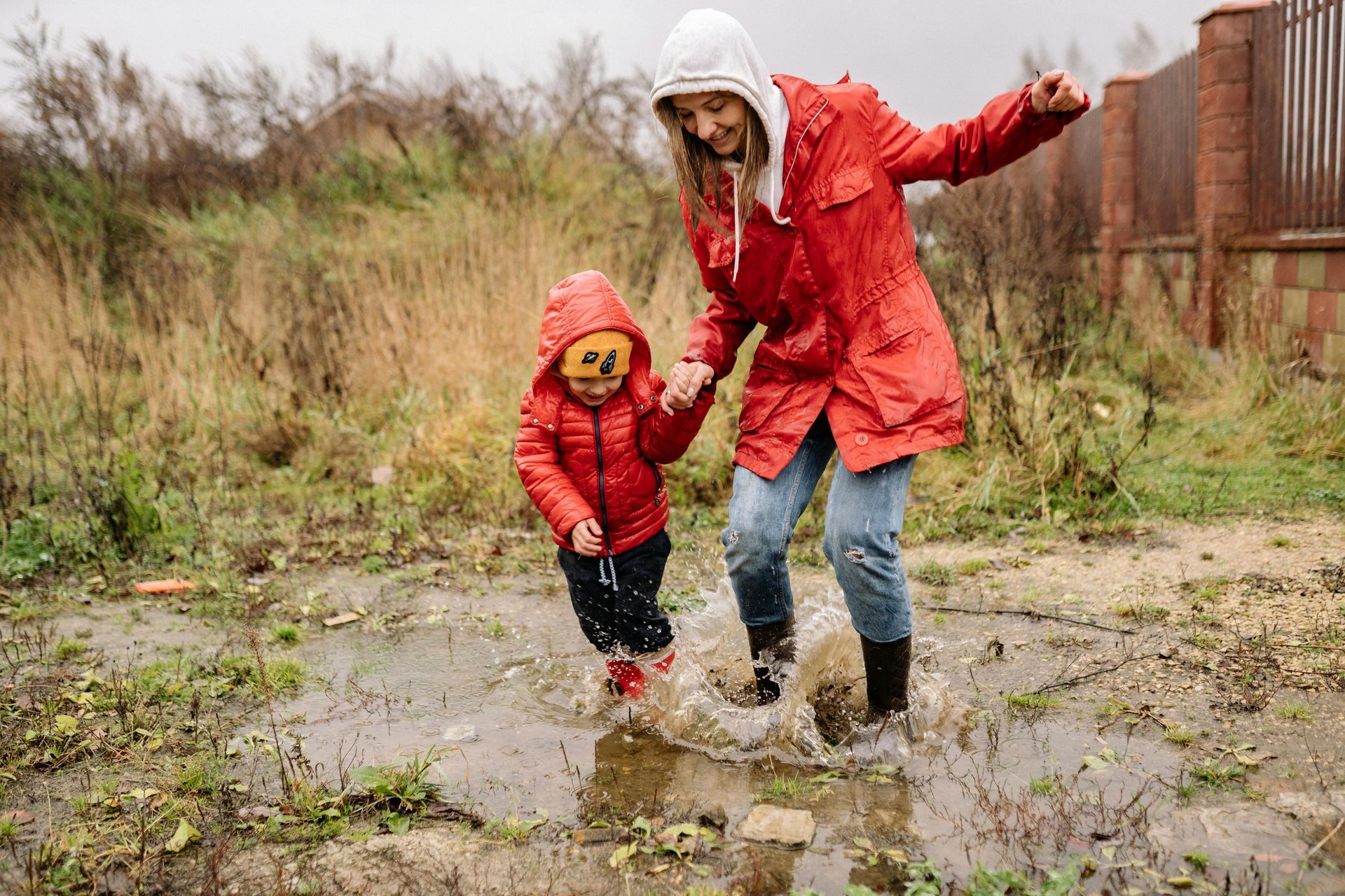 Woman and child in red raincoats jump in a puddle, smiling, on a rainy day outdoors.