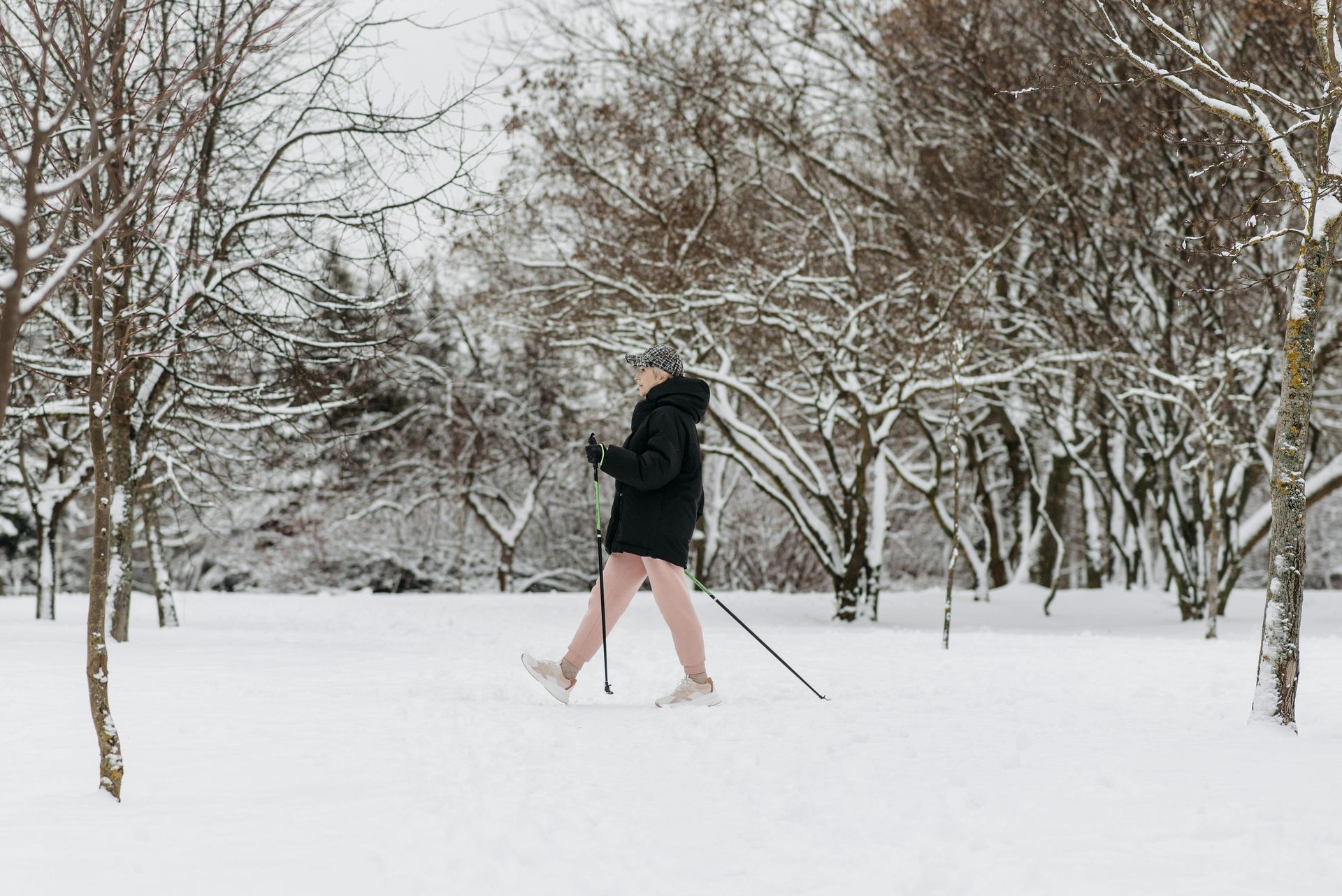 Person walking in snow-covered park, using poles for support. Trees with snow, cloudy day.
