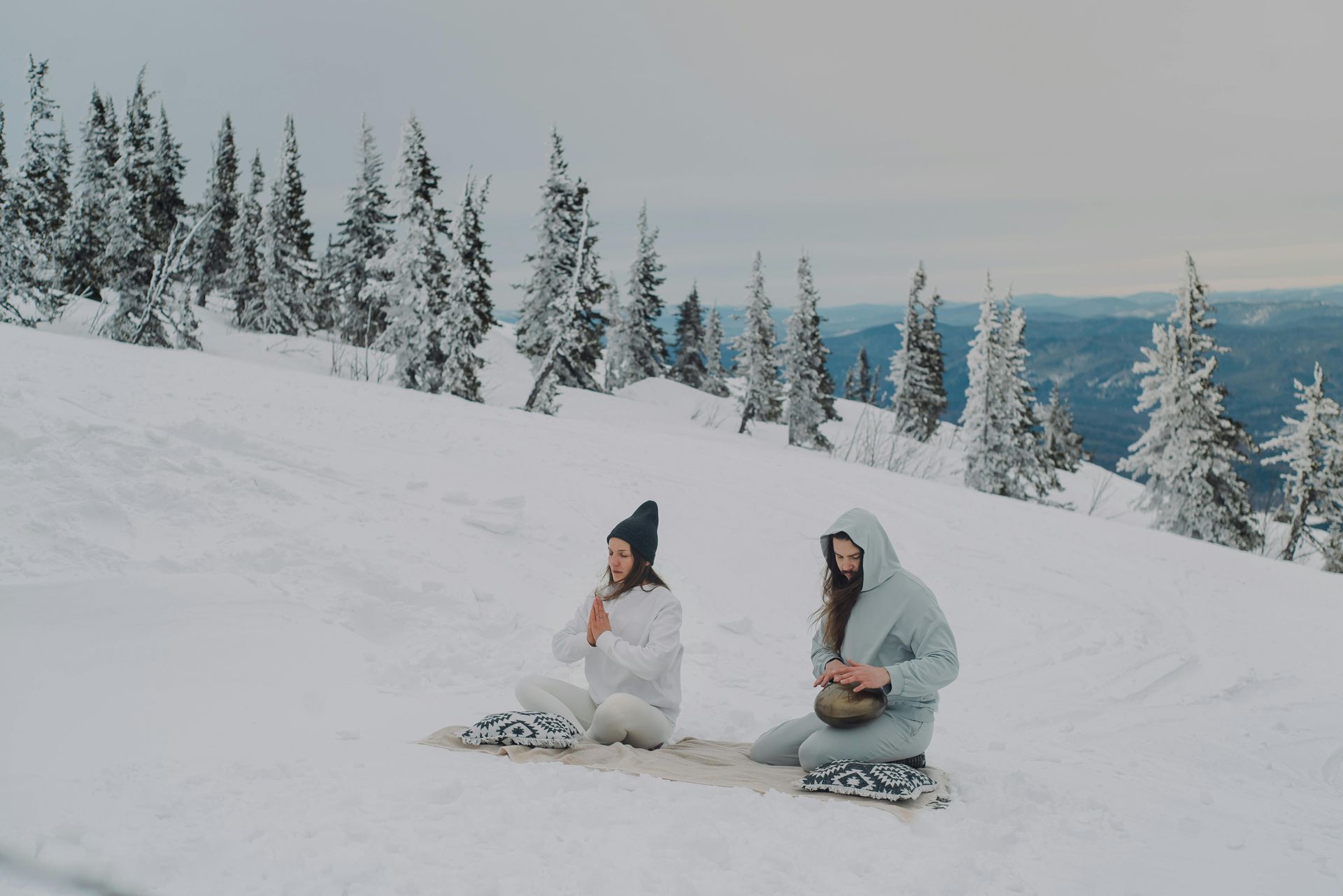 Two people sitting on snow-covered mountain, meditating. Snowy trees in background.