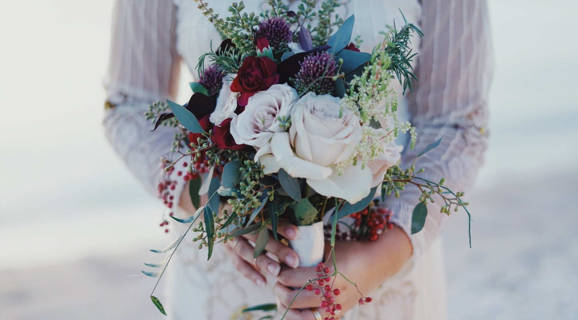 A bride holding a bridal bouquet made of white roses, red roses, and thistle