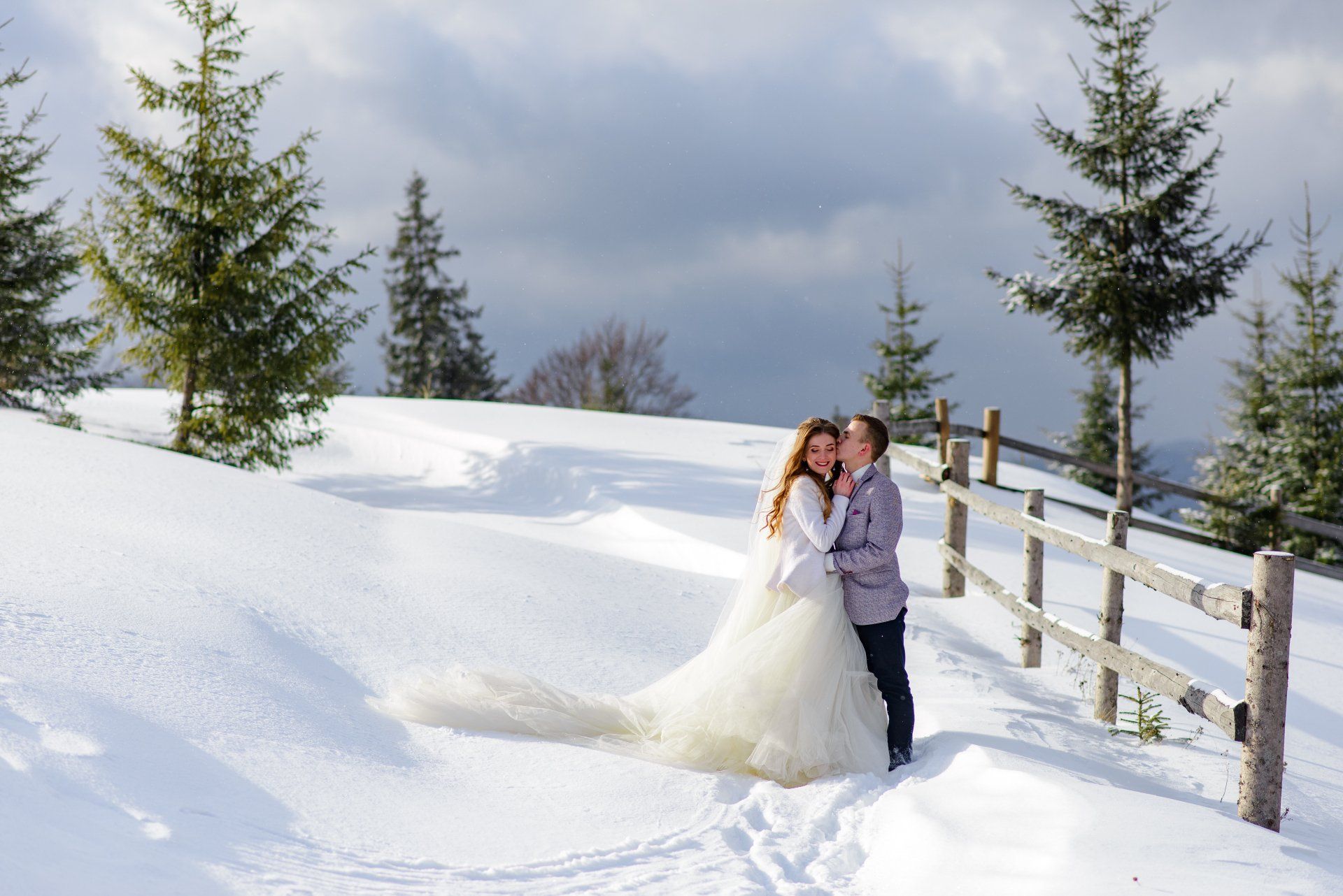 A groom kisses the bride against the backdrop of snow and trees
