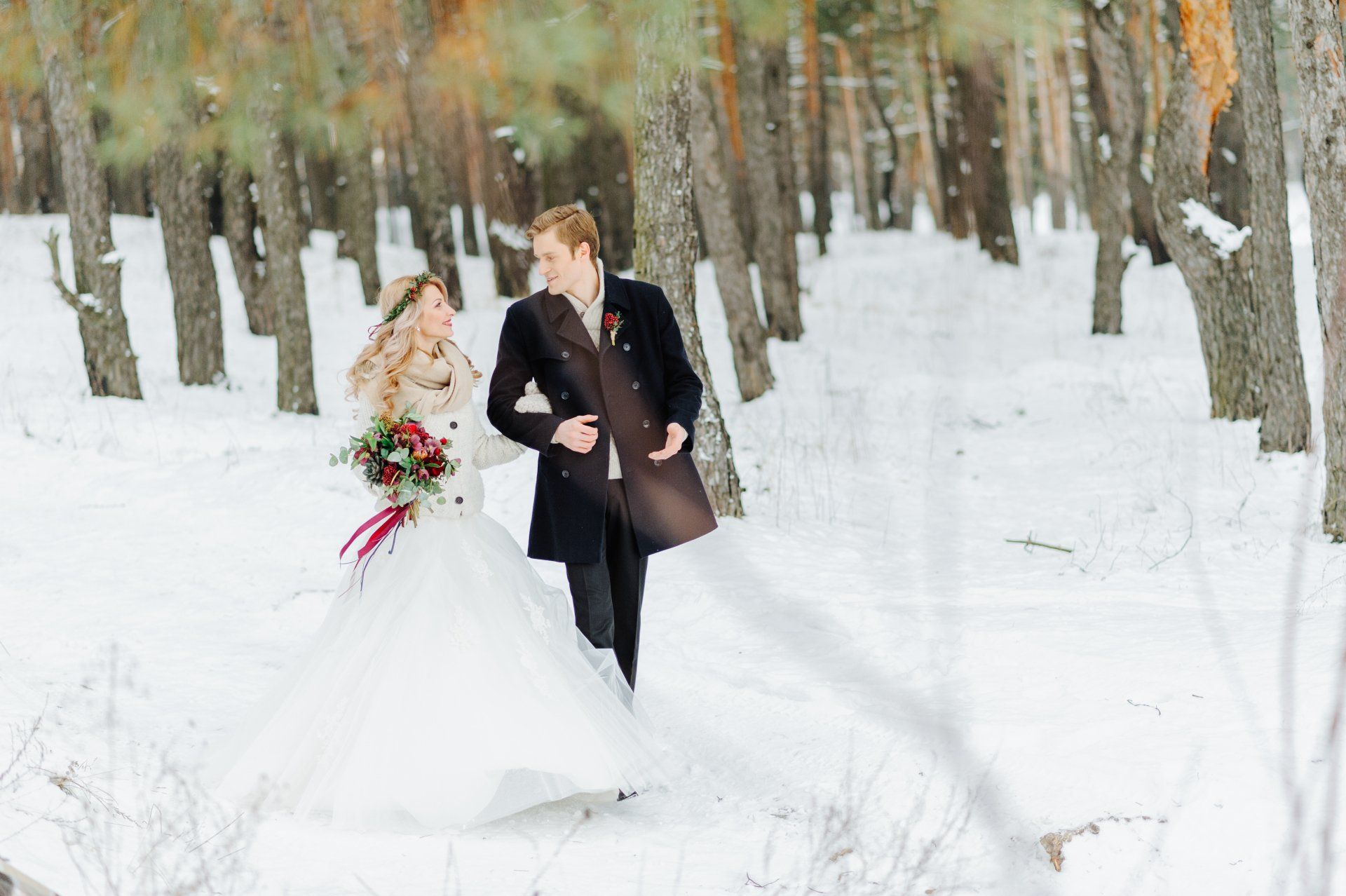 A bride and groom walk through a snow-covered forest wearing coats