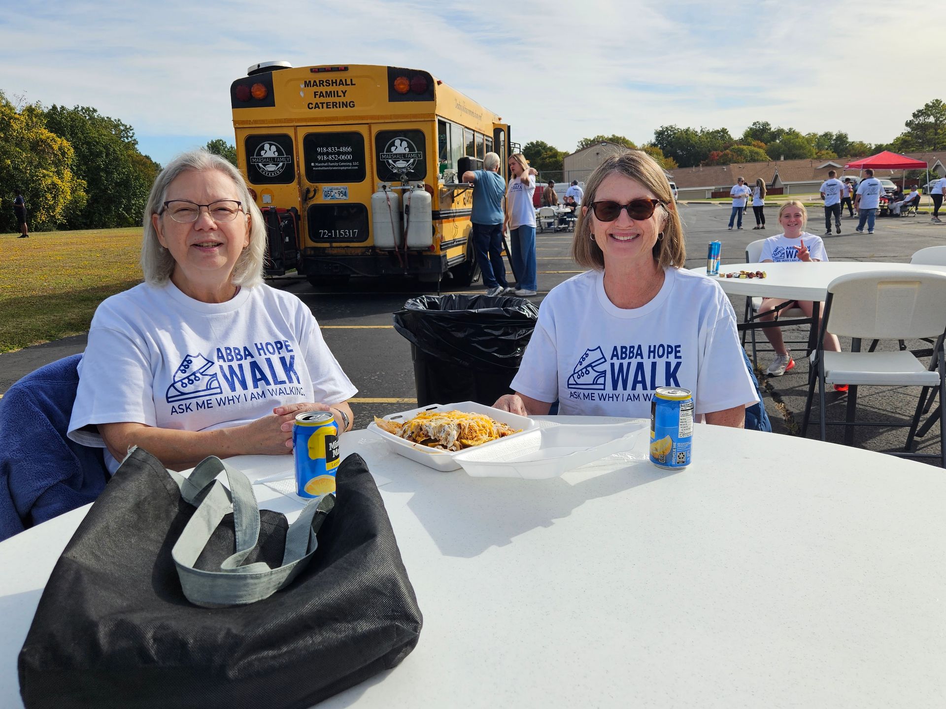 Two women are sitting at a table with food and drinks in front of a yellow school bus.