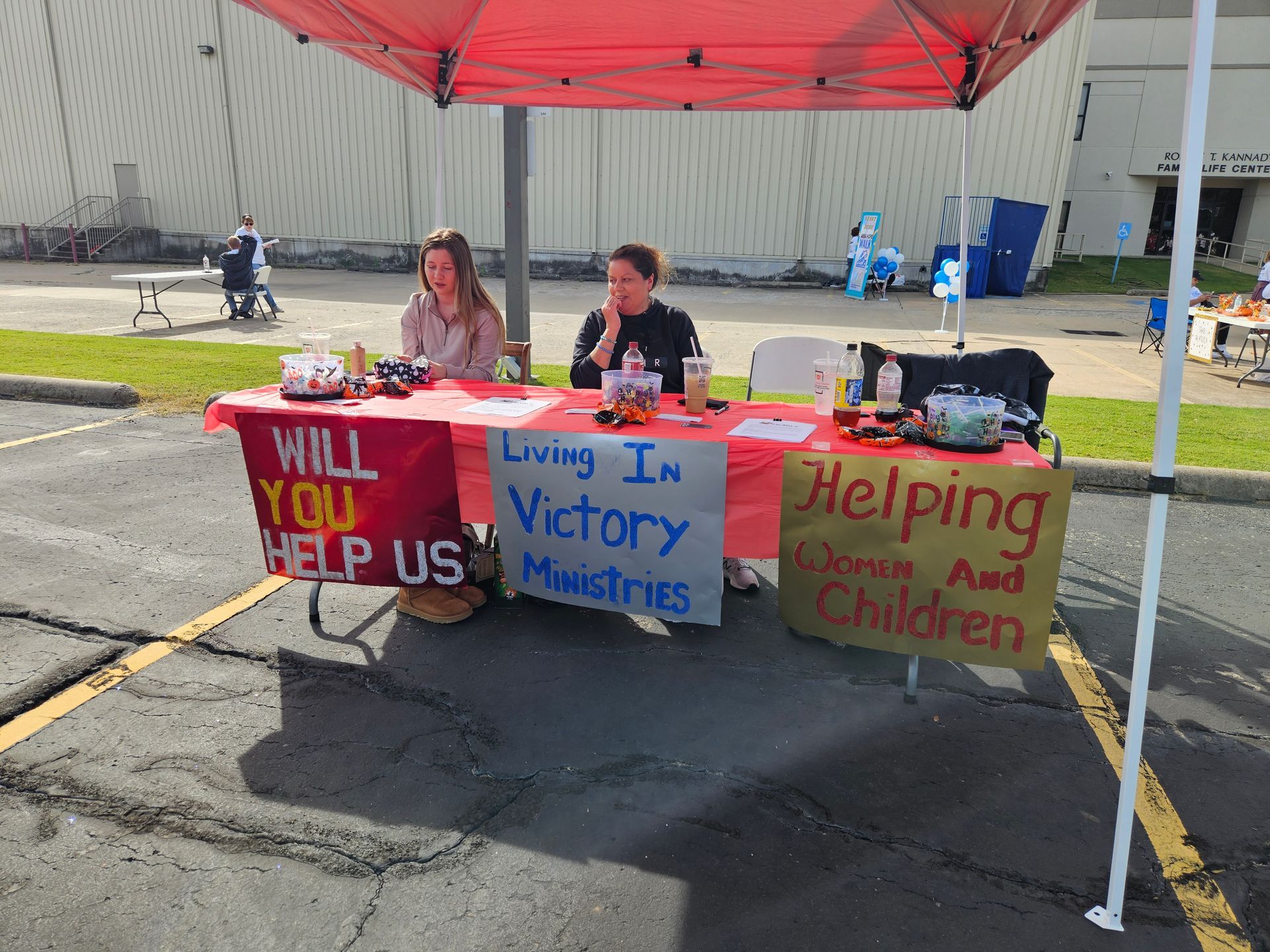 Two women are sitting at a table with a sign that says will you help us
