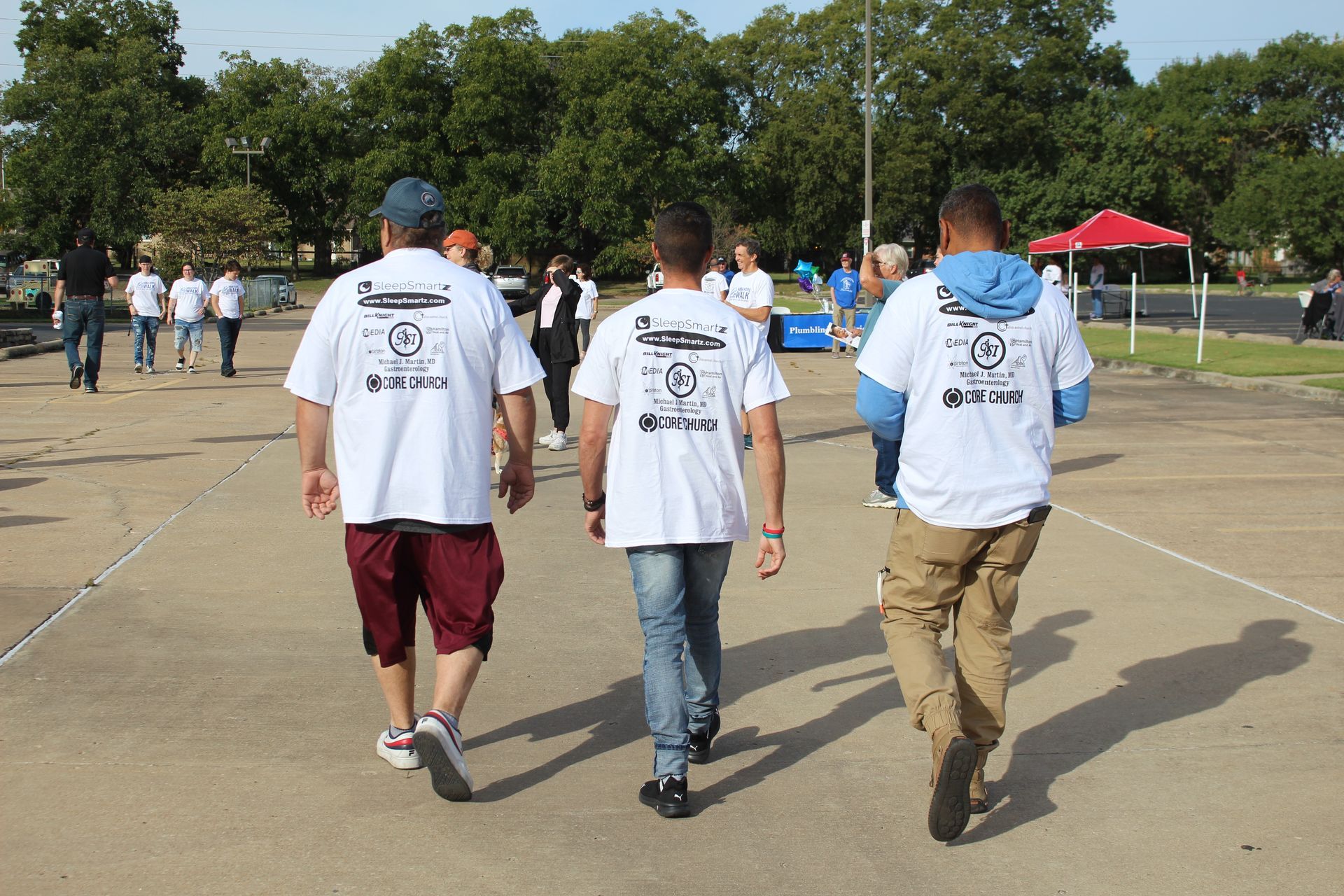 A group of men are walking down a sidewalk wearing white shirts with a logo on the back.