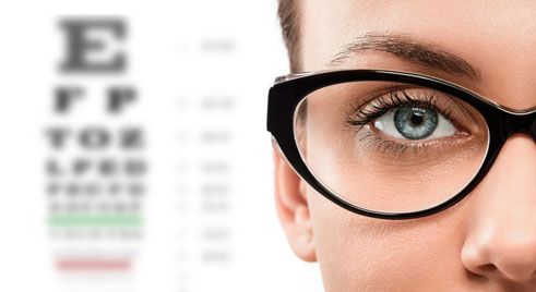 A close up of a woman wearing glasses with an eye chart in the background.