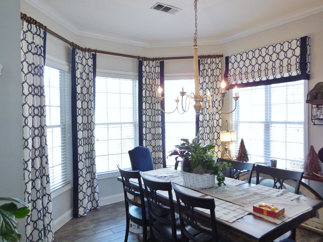 Bright dining room with large windows, patterned curtains, chandelier, and a table set with flowers.