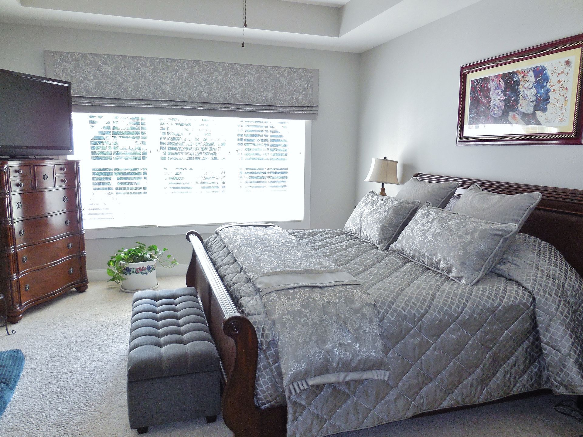 Bright bedroom with gray bedding, tufted bench, dresser, window seat, and framed wall art.