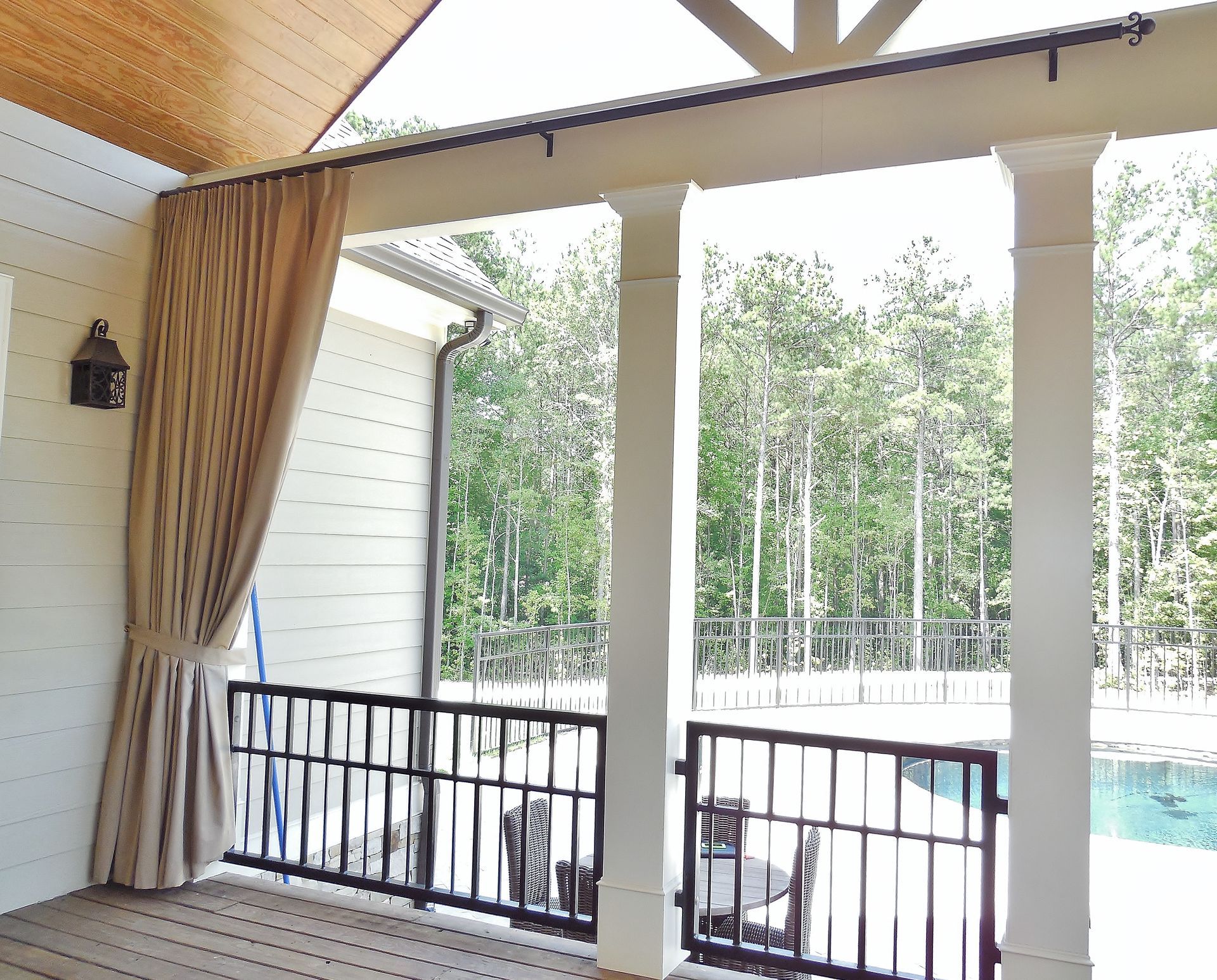 Covered patio with curtains, white columns, black railing, and a view of a pool and trees