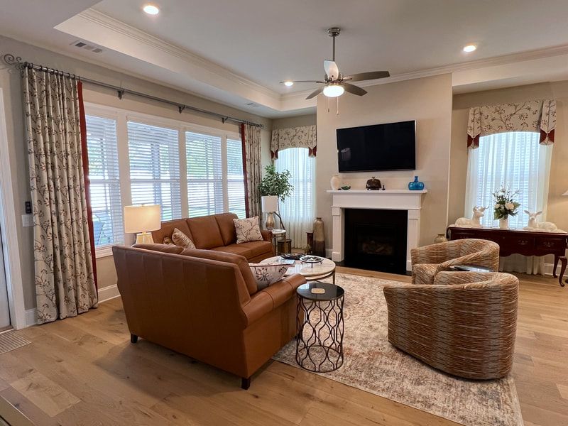 Bright living room with brown sofa, wicker chairs, fireplace, TV, and large windows with curtains.