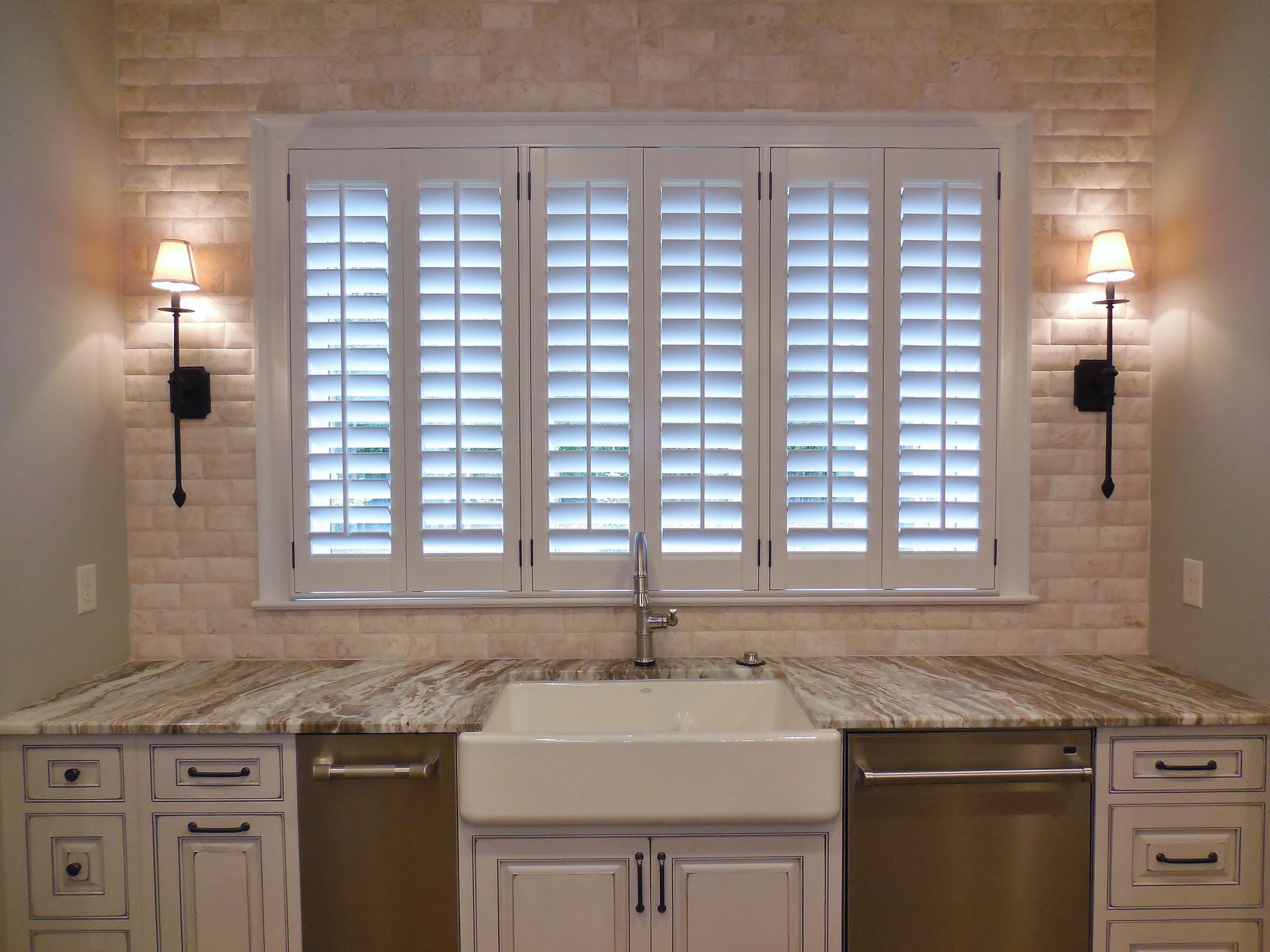 Kitchen with white window shutters, stone backsplash, sink, and wall sconces above granite countertops