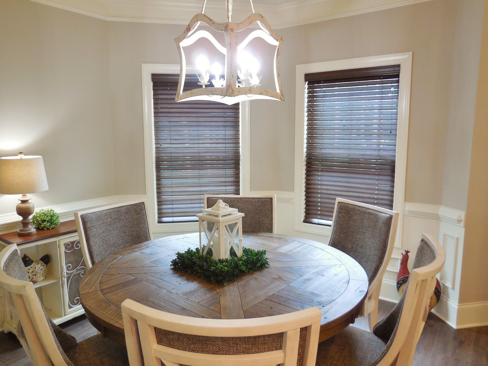 Round wooden dining table in a beige dining room with upholstered chairs, windows, and a hanging light fixture.