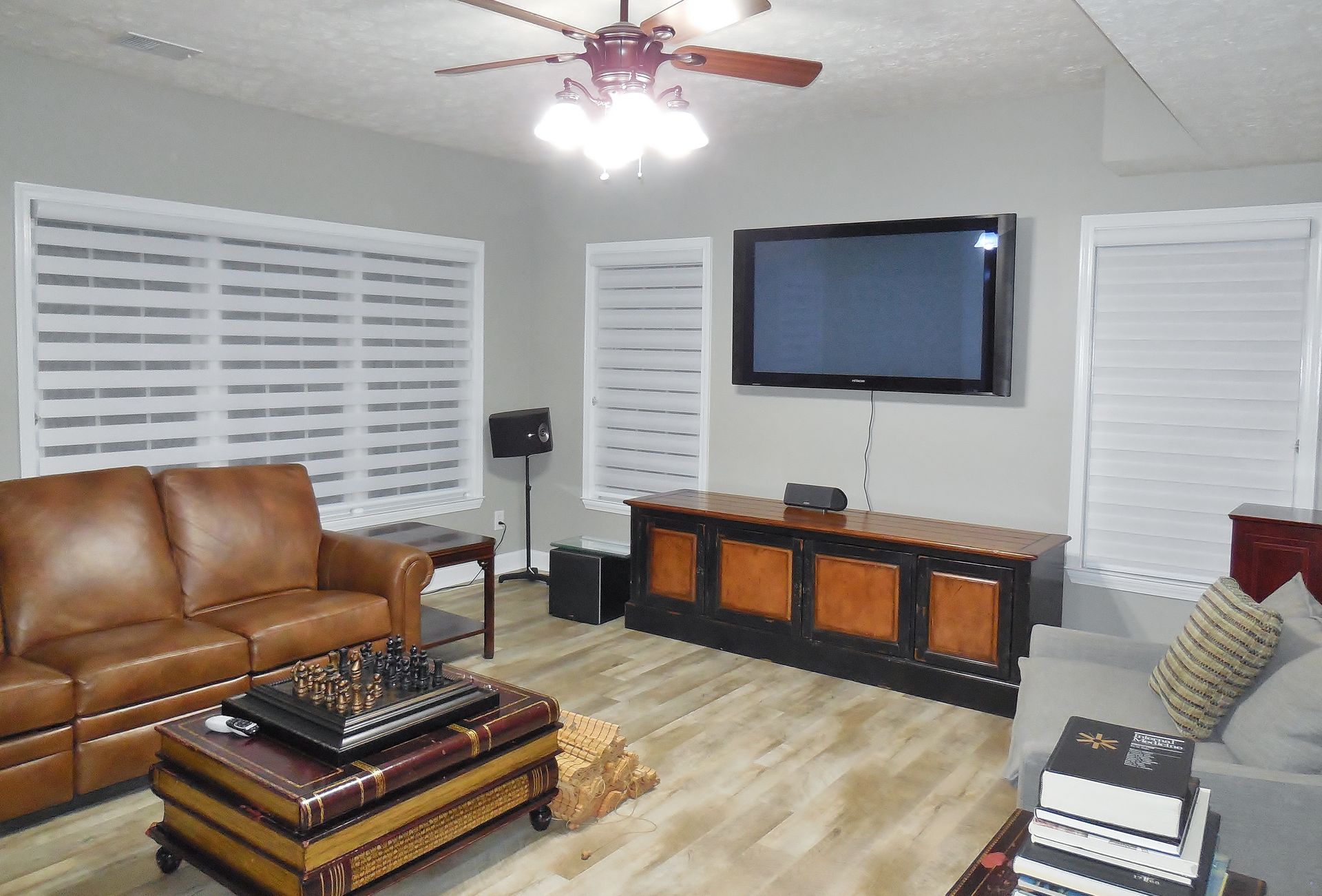 Living room with brown leather sofa, wall-mounted TV, white shutters, and ceiling fan