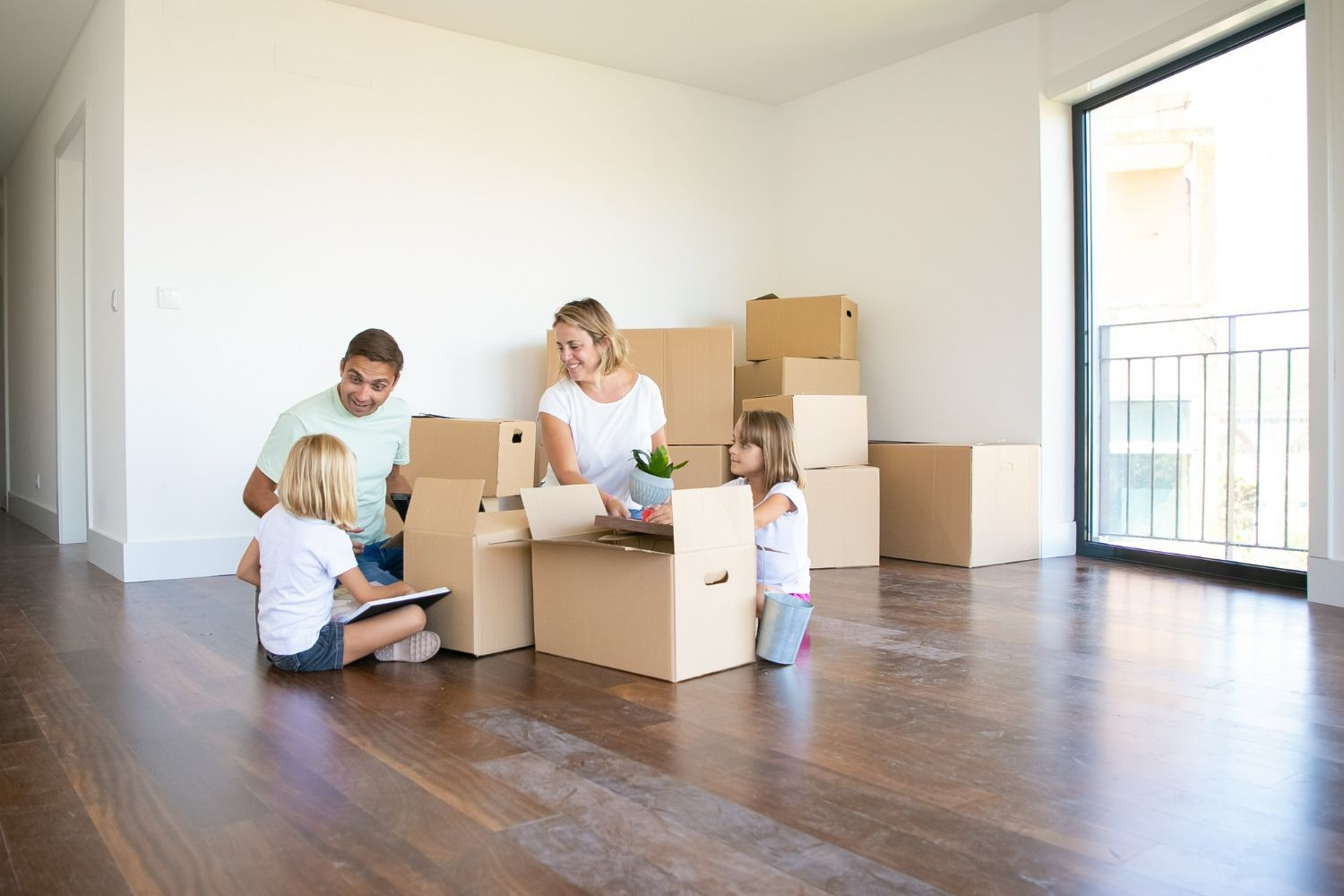 A family is sitting on the floor in an empty room surrounded by cardboard boxes.