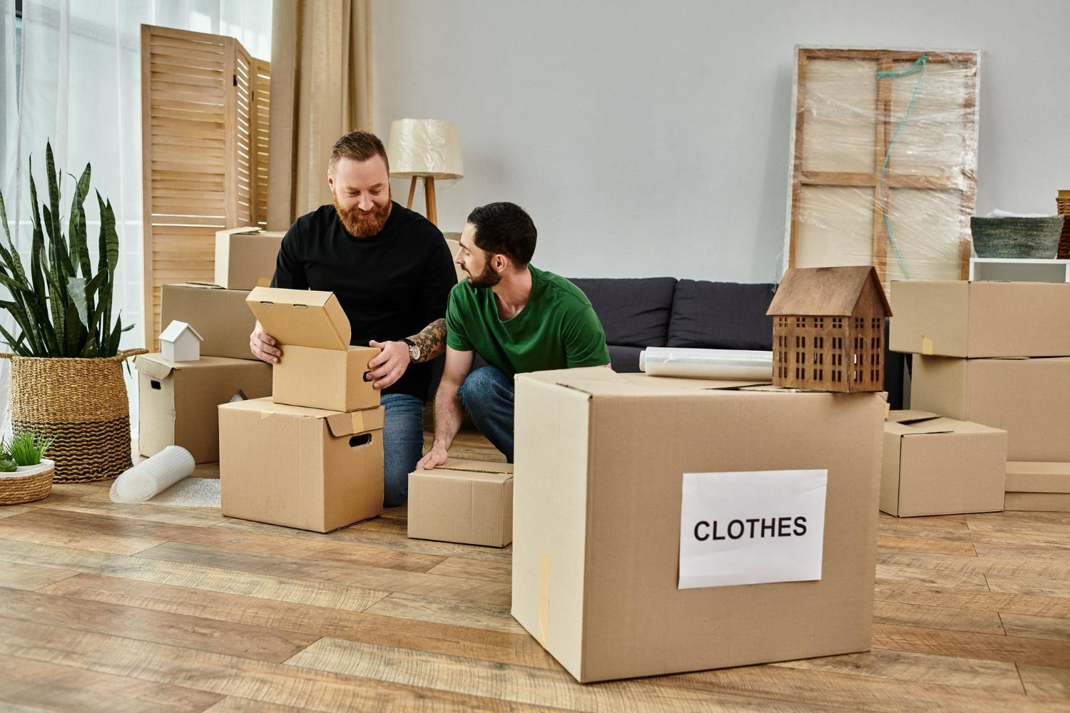 Two men are sitting on the floor in a living room surrounded by cardboard boxes.