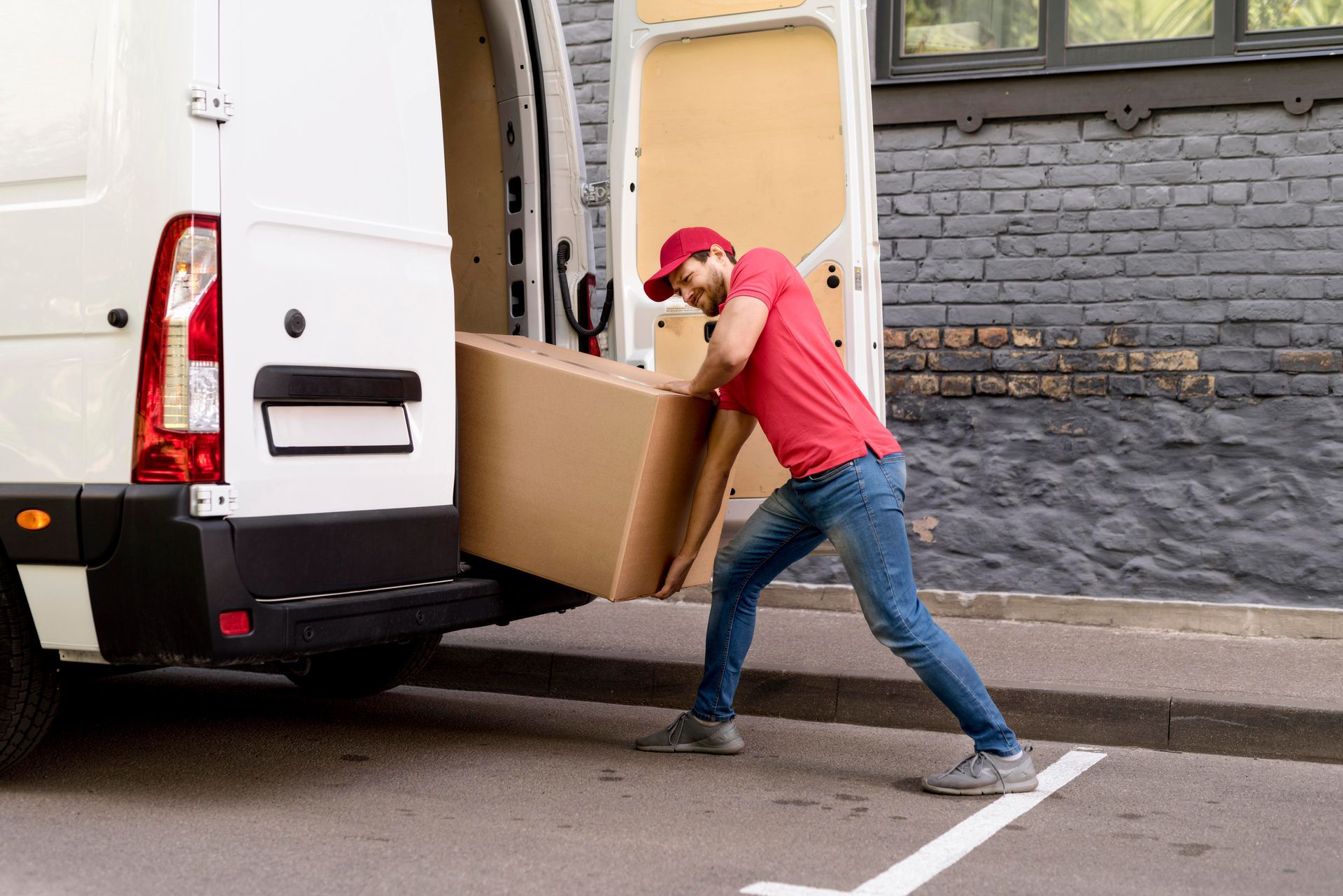 Delivery person in red shirt and cap, pulling a large cardboard box from the back of a white van on a city street.