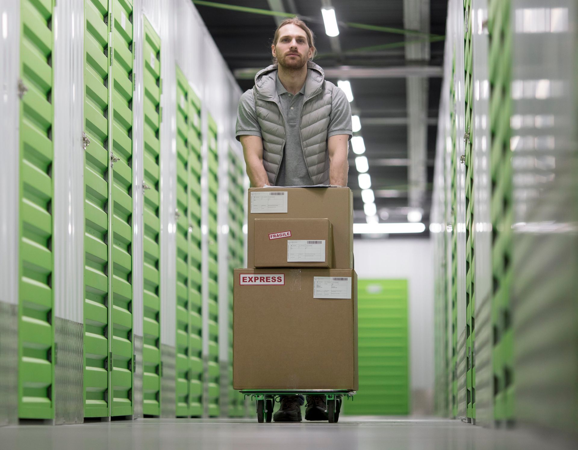 Man in storage facility pushing boxes on a dolly between green lockers.