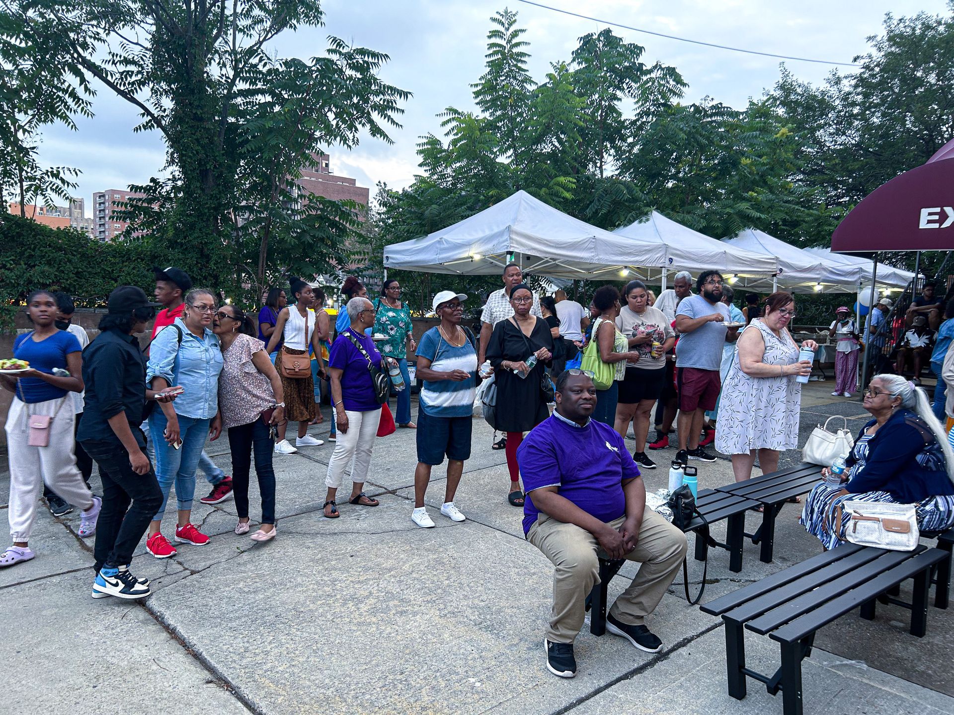 A group of people are standing and sitting on benches in a park.
