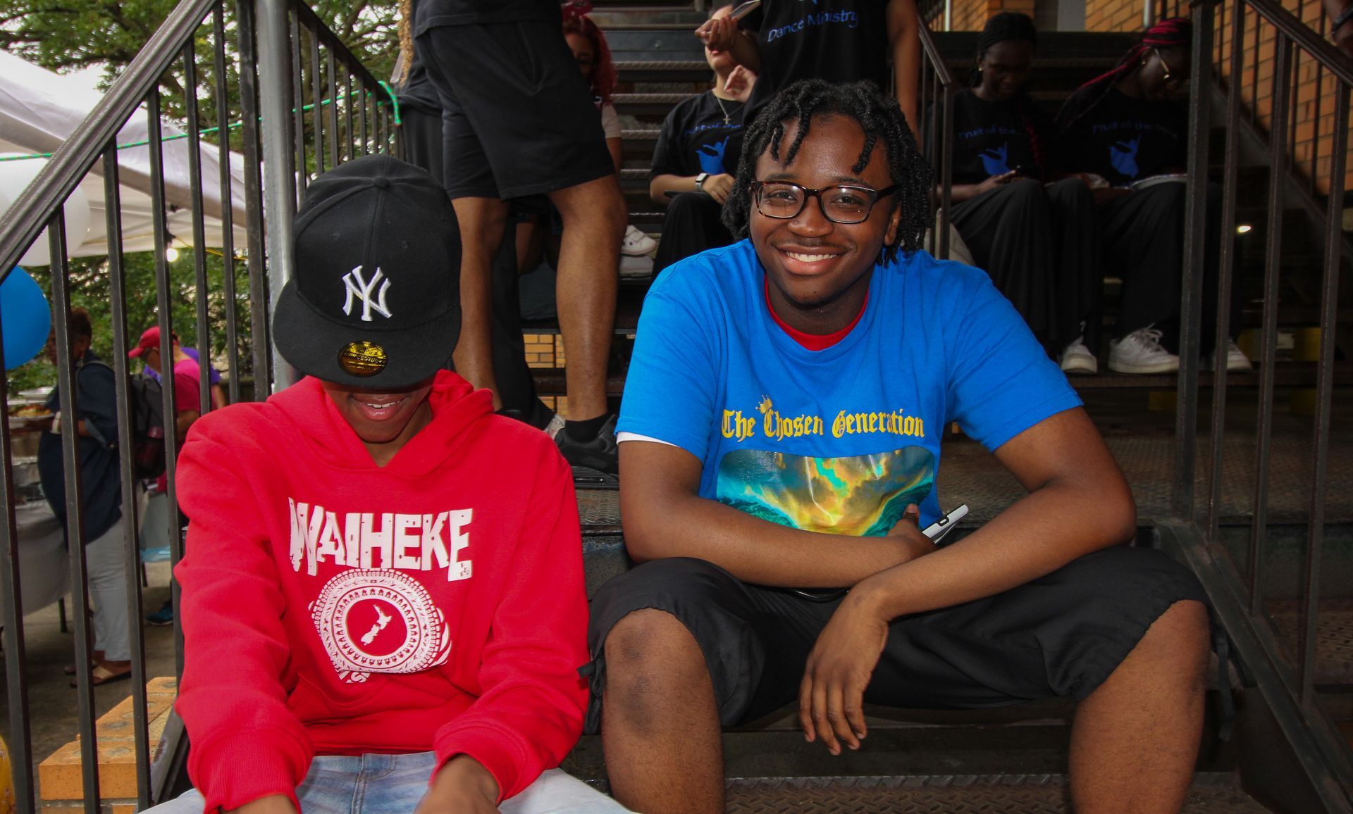 Two young men are sitting on a set of stairs one is wearing a blue shirt and the other is wearing a red hoodie