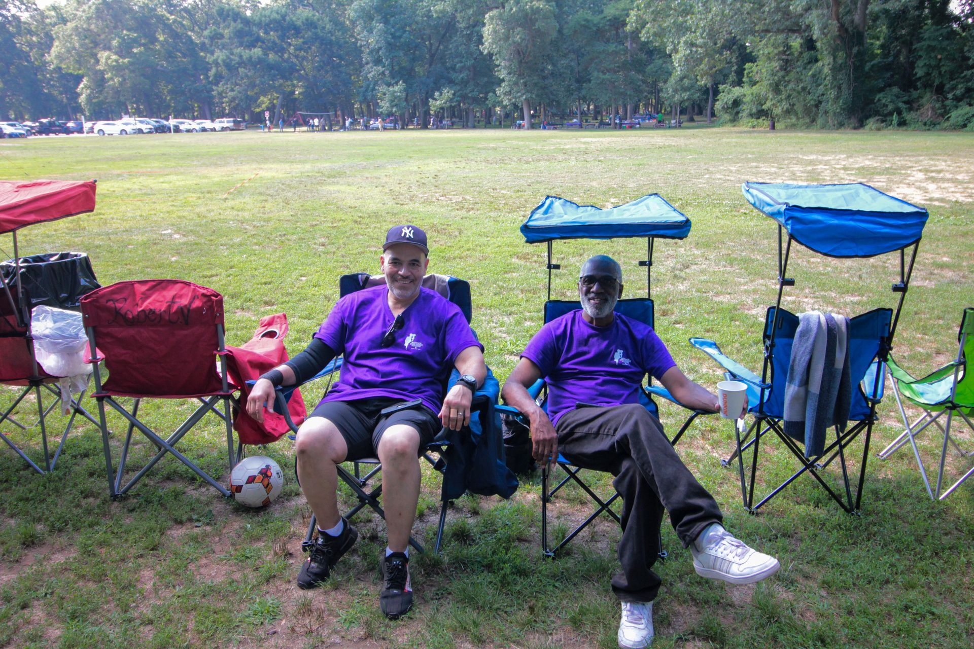Two men in purple shirts are sitting in folding chairs in a field.
