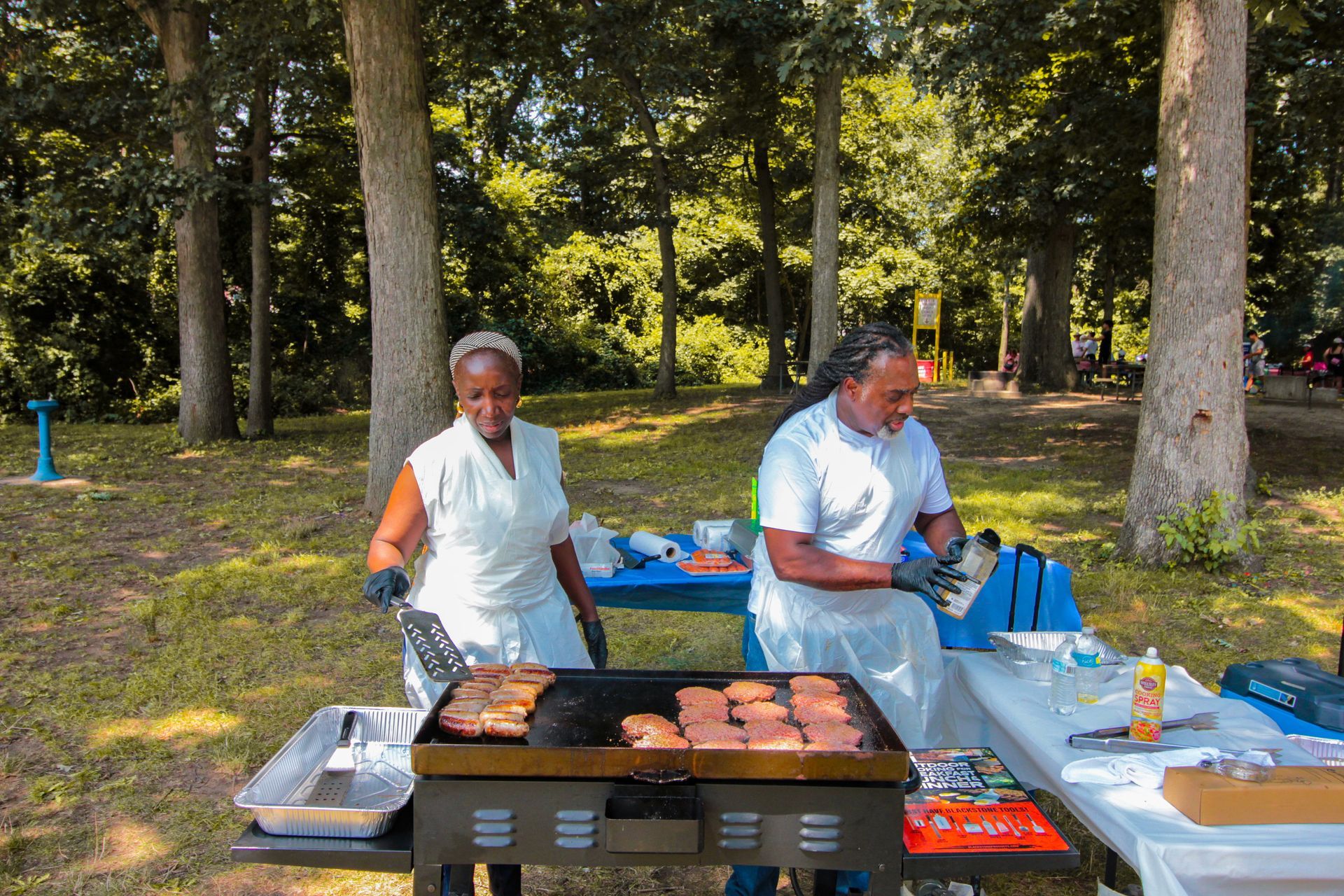 Two people are cooking food on a grill in a park.
