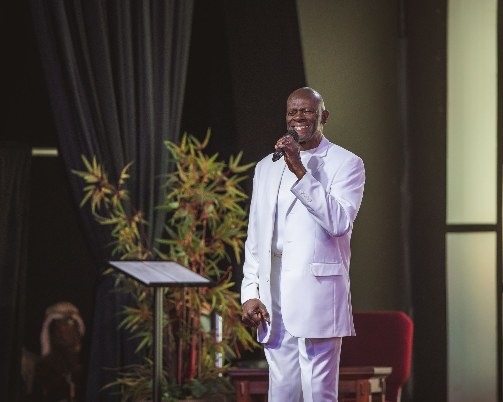 A man in a white suit is standing in front of a podium holding a microphone.