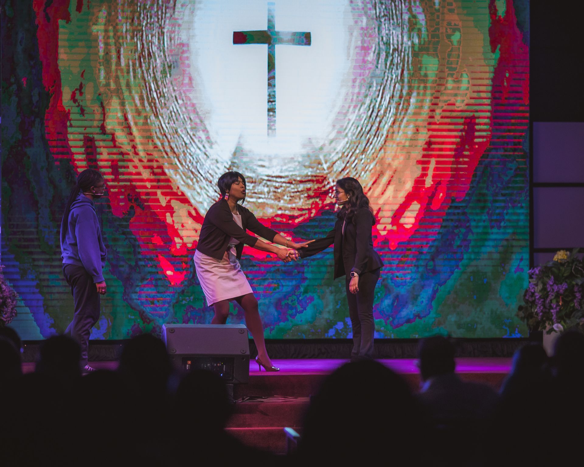 Two women are shaking hands on a stage in front of a large screen with a cross on it.