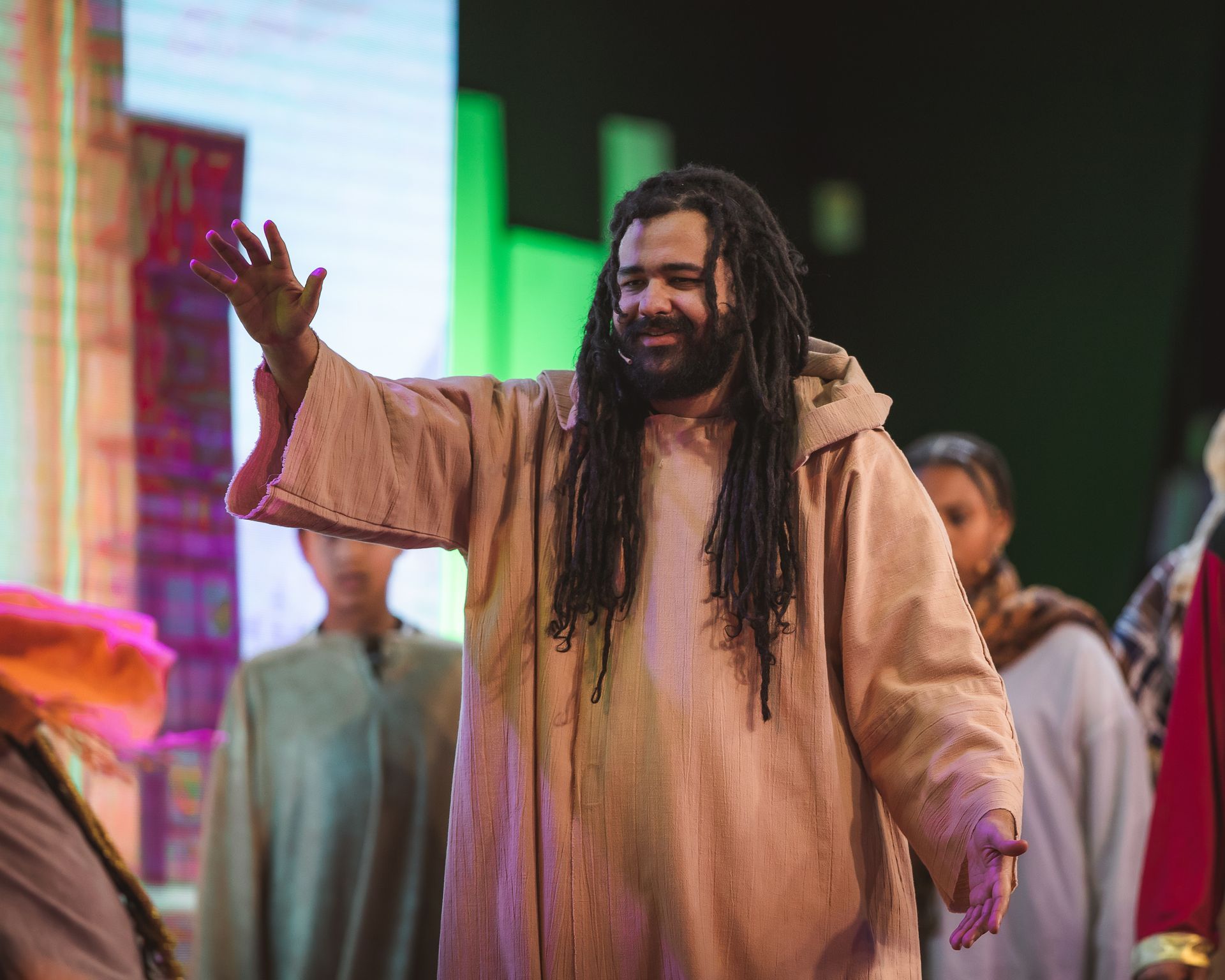 A man with dreadlocks is dressed as jesus and is waving his hand.
