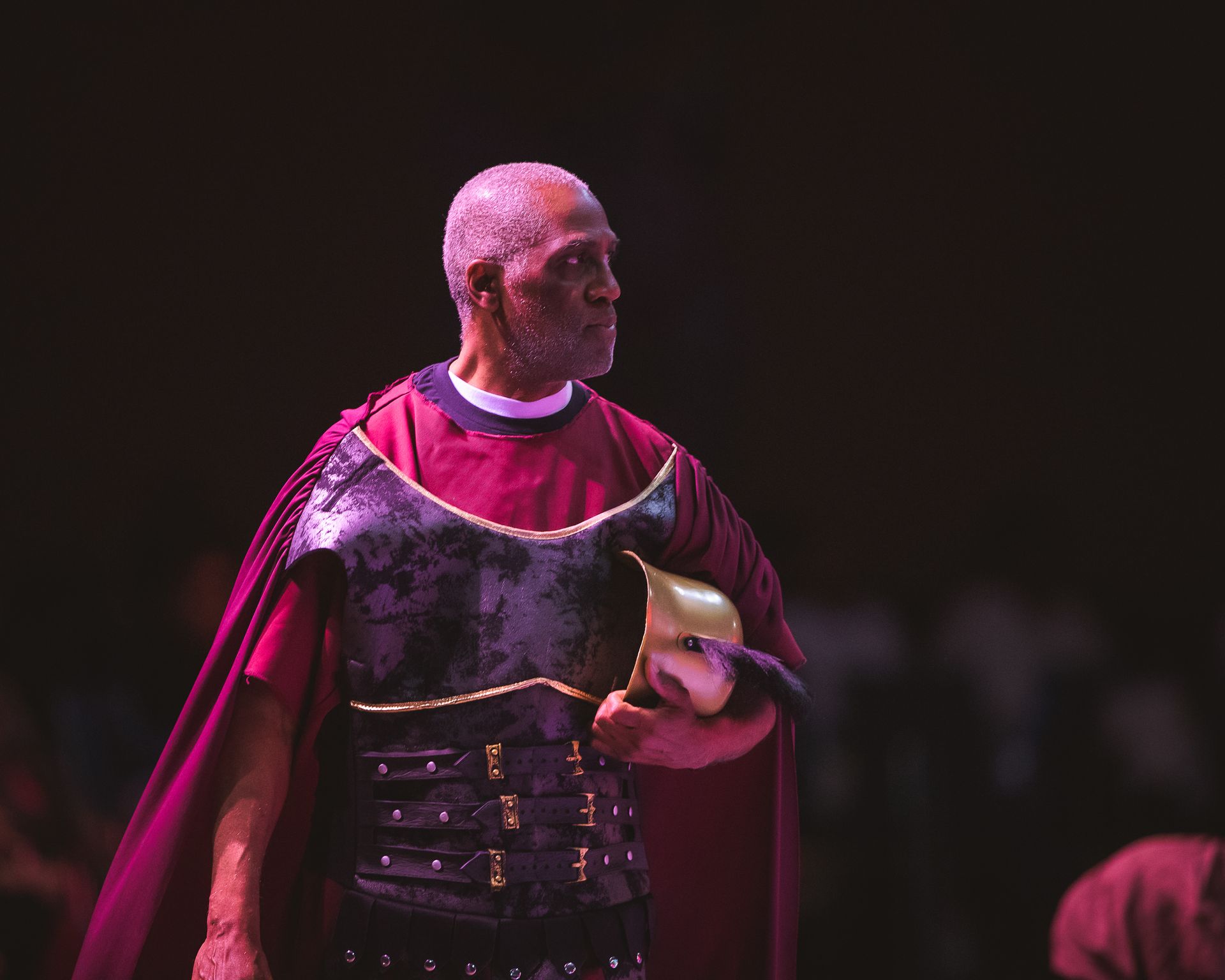 A man in armor is holding a mask on a stage.