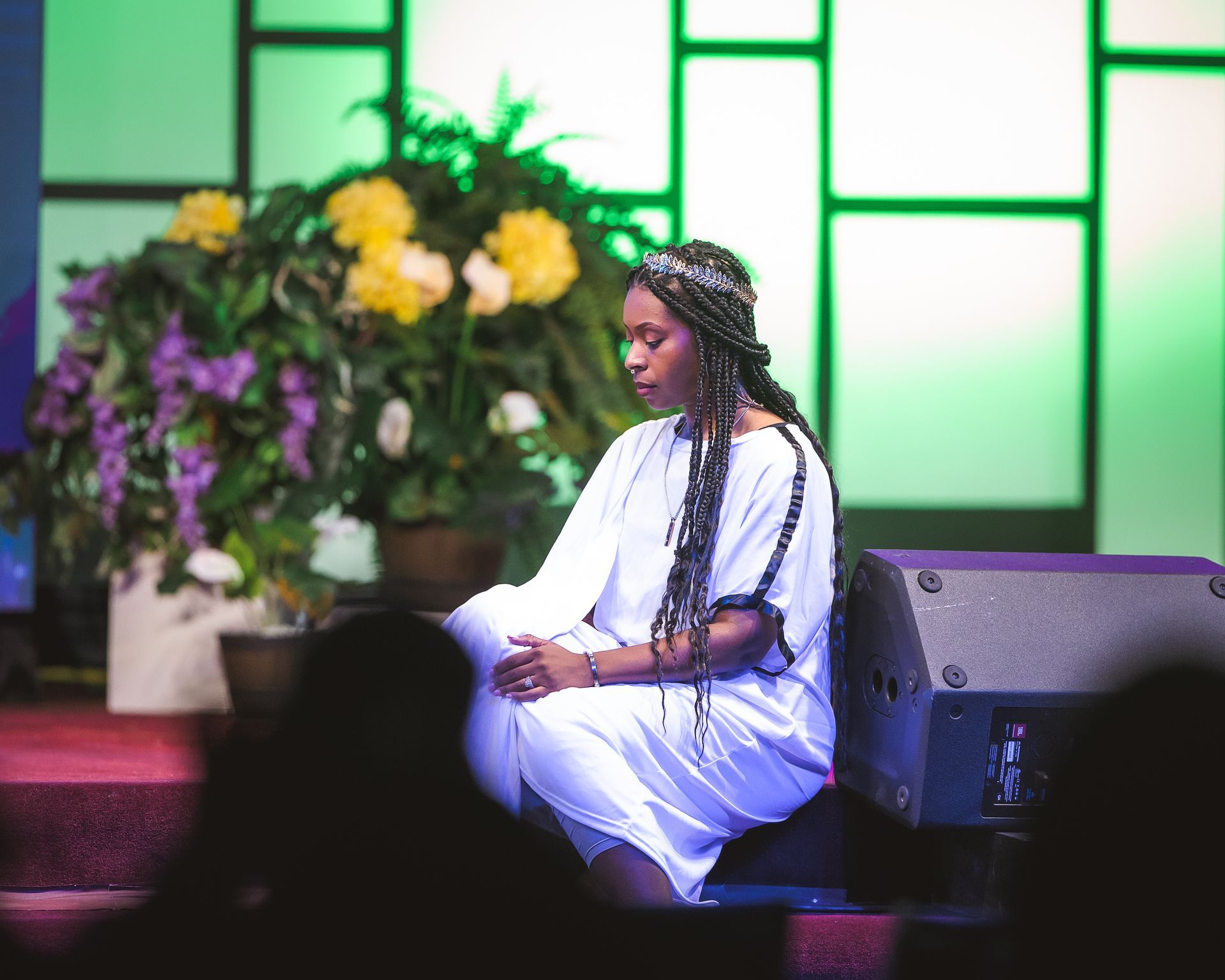 A woman in a white dress is sitting on a stage in front of a speaker.