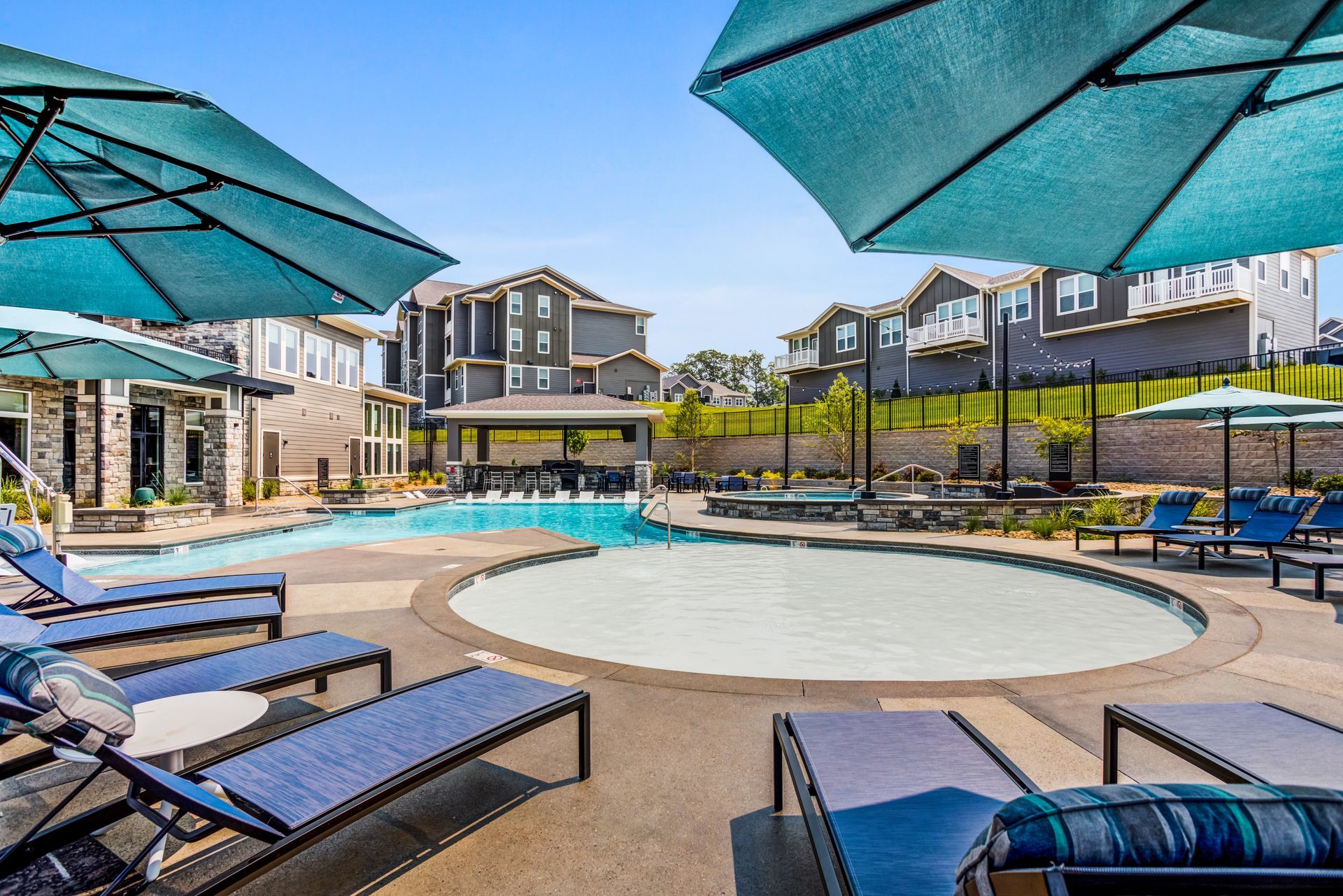 Pool area with lounge chairs, umbrellas, and buildings in the background on a sunny day.