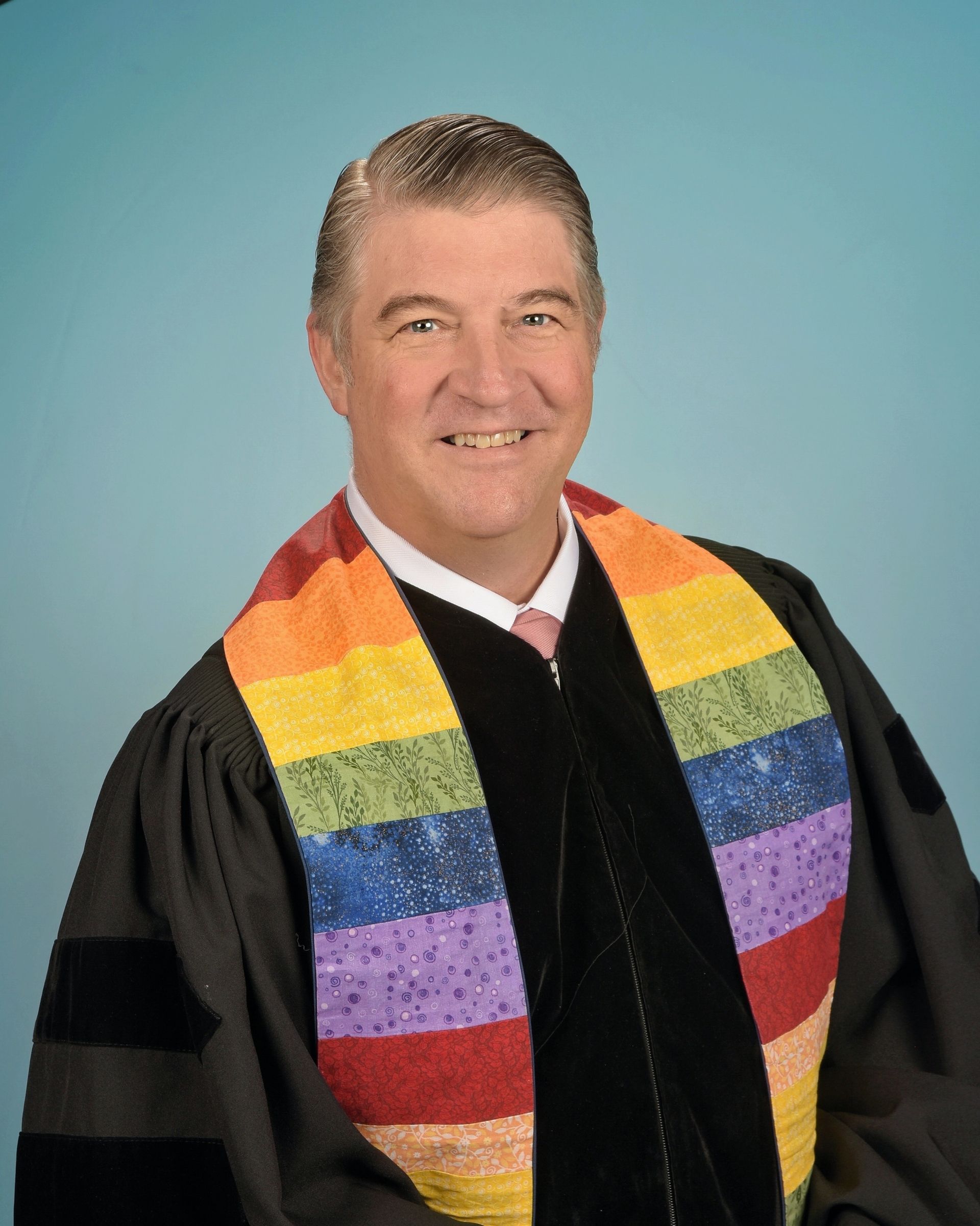 Man with glasses and rainbow stole smiles, wearing clergy robes.