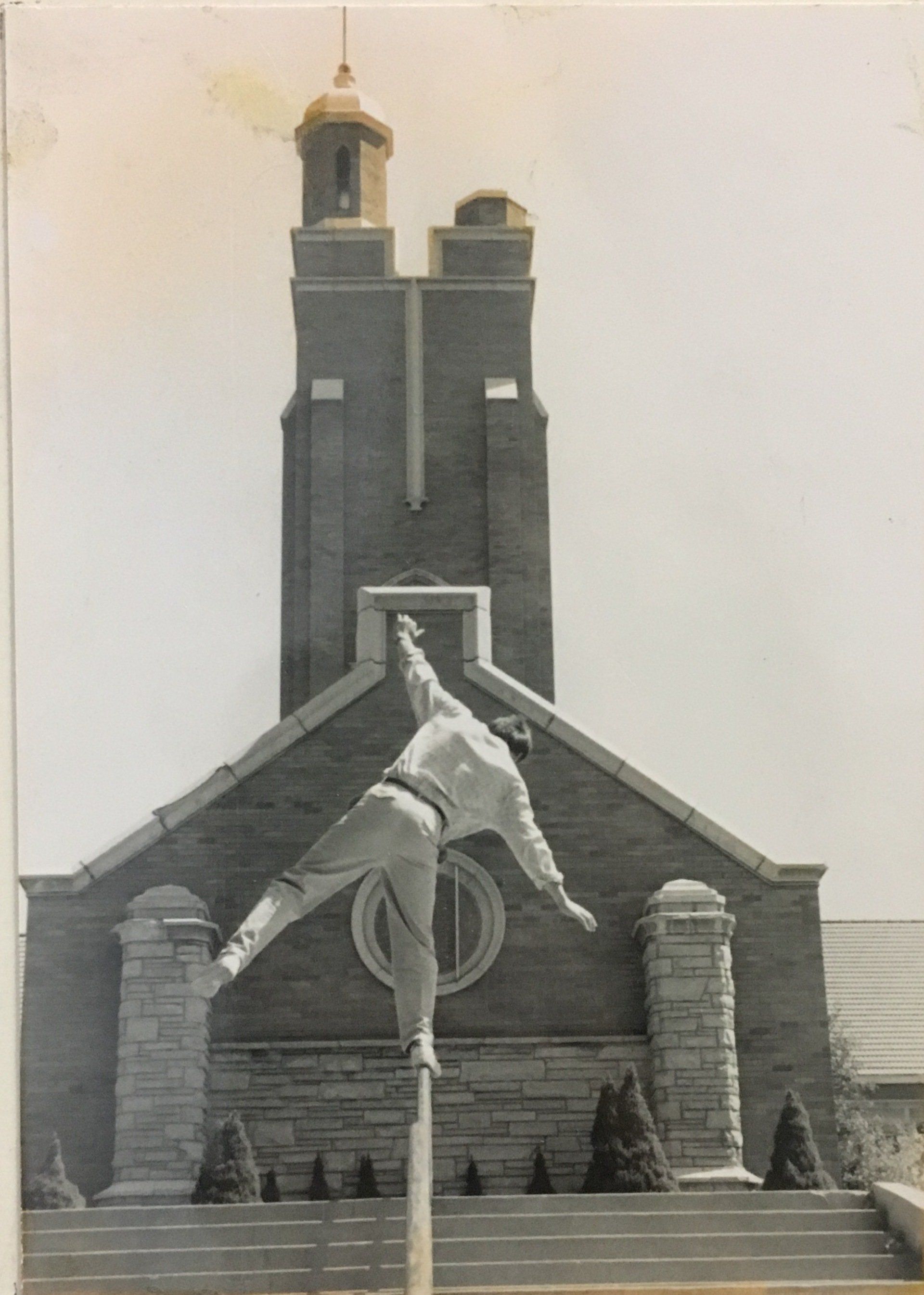 A person balances on a pole in front of a brick building with a steeple.