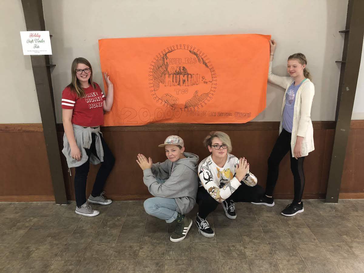 Group of five kids holding up an orange banner with a design. They are clapping and smiling in front of a wall.