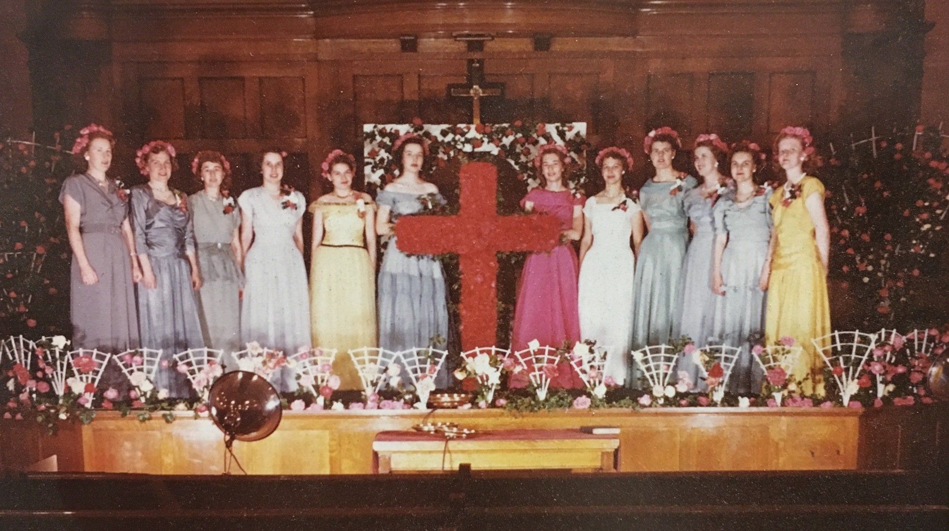 Group of women in gowns posing behind a large red cross on stage, decorated with flowers.