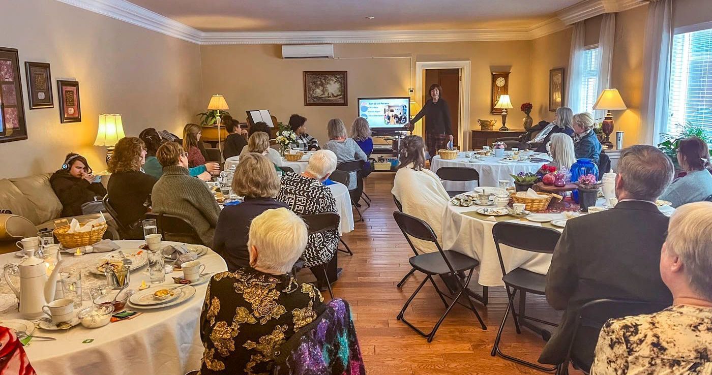 People in a room seated at tables, listening to a presentation. The room is set up for an event.