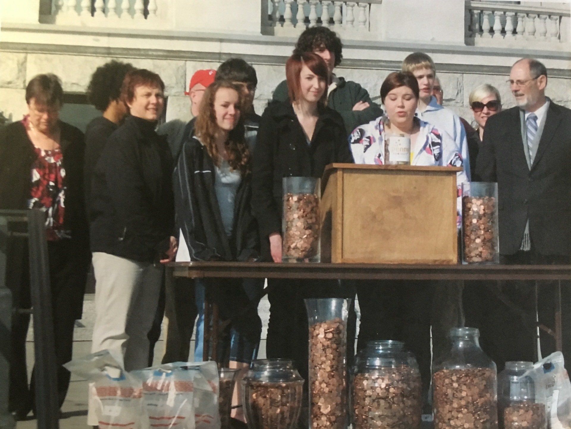Group of people at outdoor event with jars of pennies on a table; woman speaks at a podium.