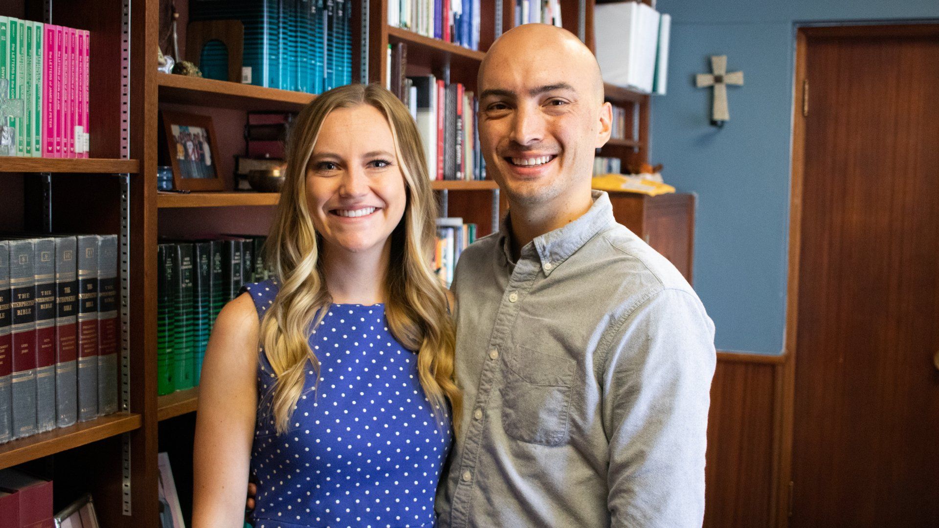 Smiling couple posing. The woman wears a blue polka-dot dress. The man has a button-down shirt.