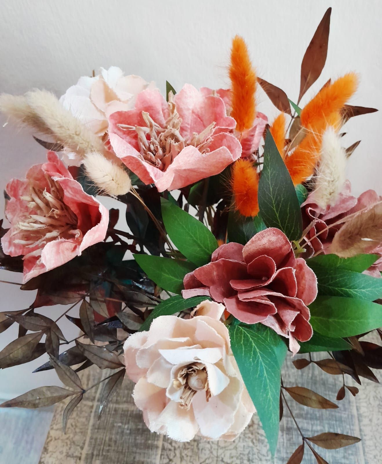 Ramo con flores de color rosa, durazno y óxido, hojas verdes y suaves detalles naranjas sobre un fondo rústico.