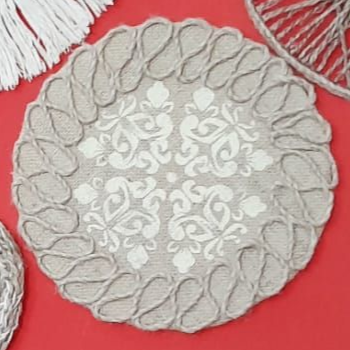 Posavasos redondo de arpillera con diseño floral blanco, borde de cuerda, sobre superficie roja.