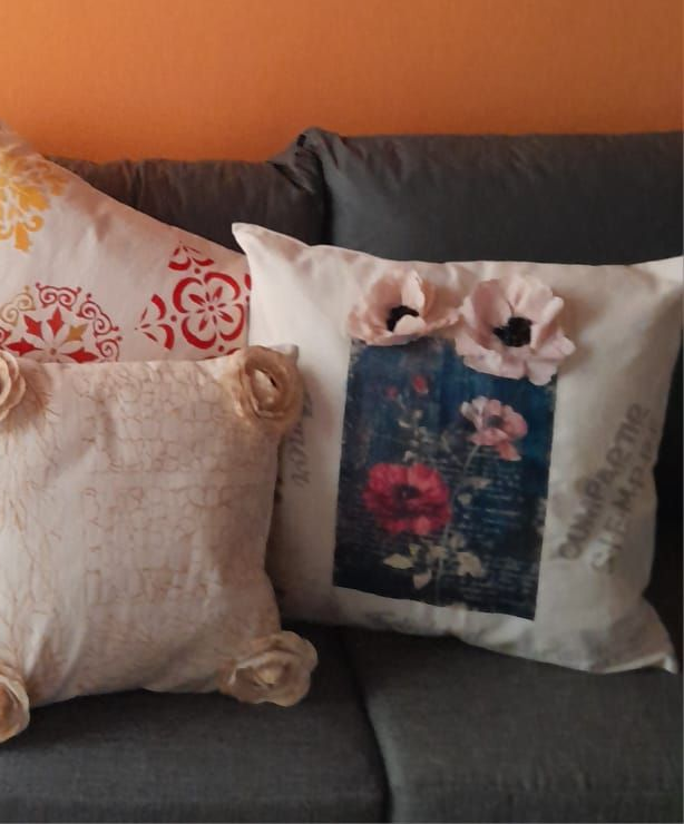 Tres almohadas decorativas sobre un sofá gris: una con estampado floral, dos con adornos de flores de tela, contra una pared