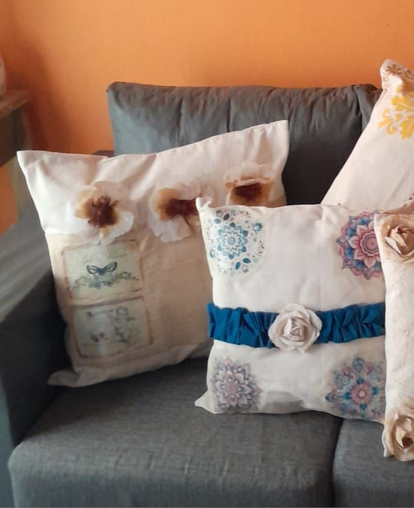 Sofá con cojines decorativos color crema. Cada uno tiene adornos: flores, patrones estarcidos y un lazo azul.