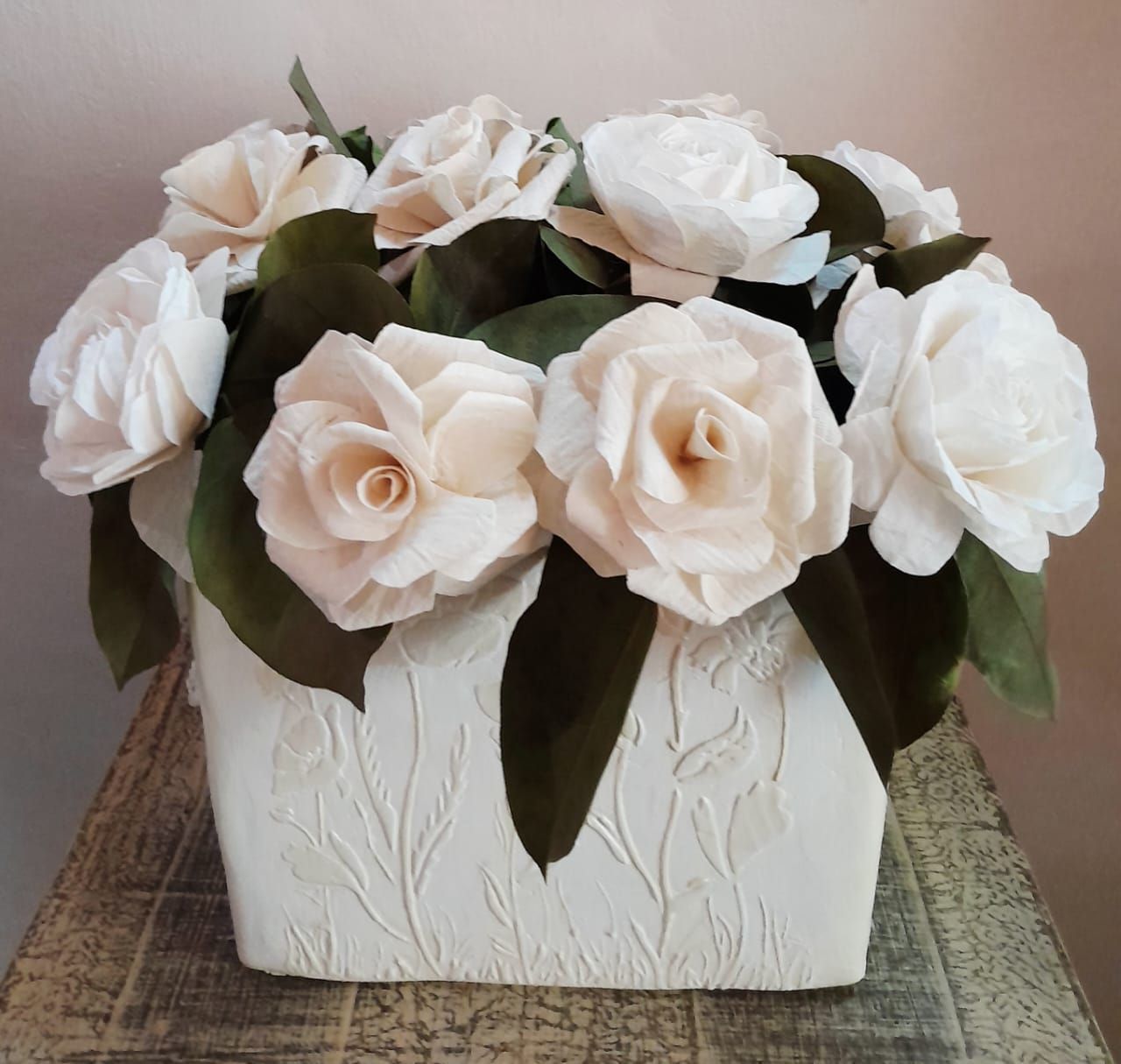 Flores blancas en un jarrón cuadrado de color crema con detalles florales en relieve, sobre una superficie de madera.