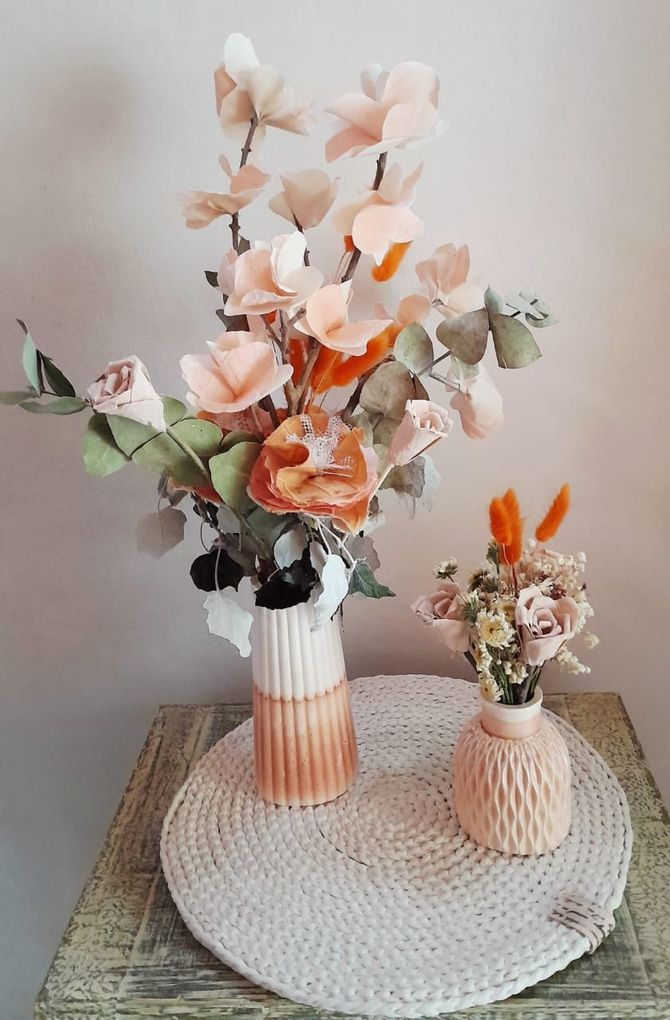 Arreglo floral en jarrones de color durazno y naranja sobre un mantel tejido encima de una mesa de madera desgastada.