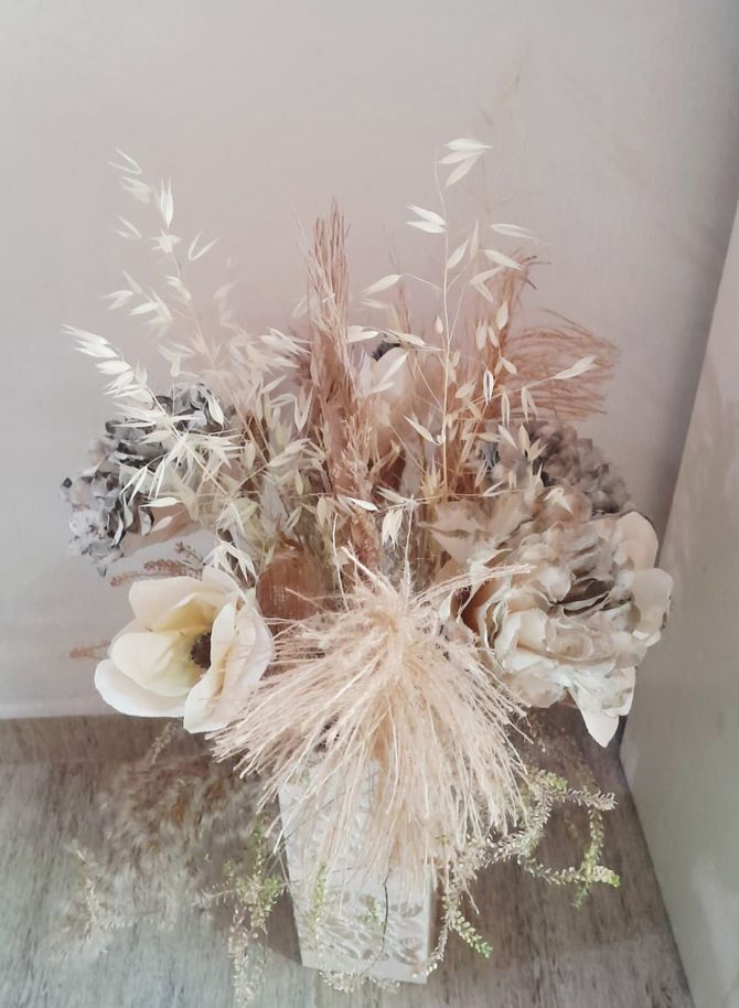 Arreglo de flores secas en un jarrón de textura transparente, con tonos neutros de crema, bronceado y marrón.
